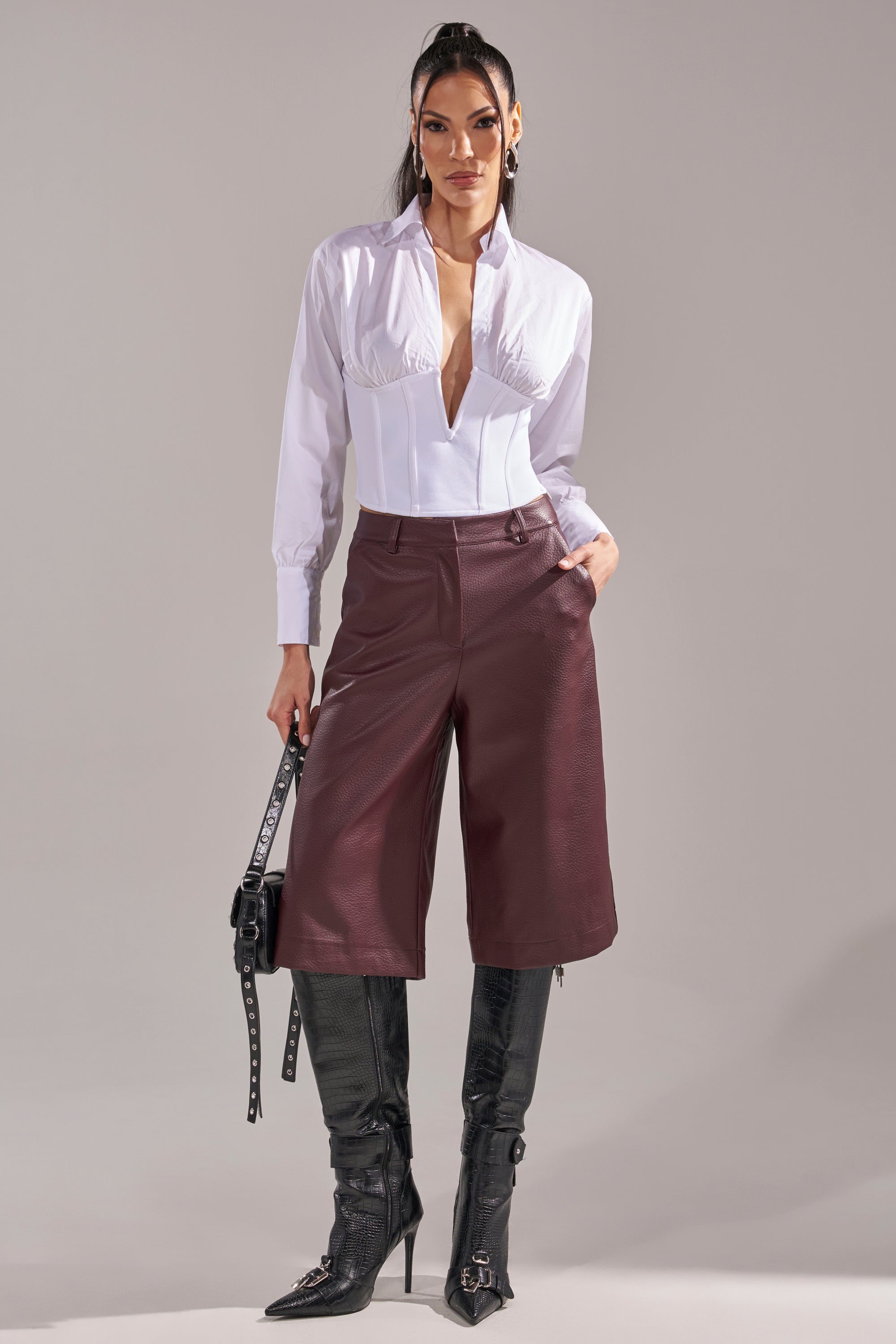 A woman stands against a plain background wearing the SNATCHED UP CORSET TOP, burgundy knee-length shorts, tall black boots, and holding a black handbag with her hair styled back. 5
