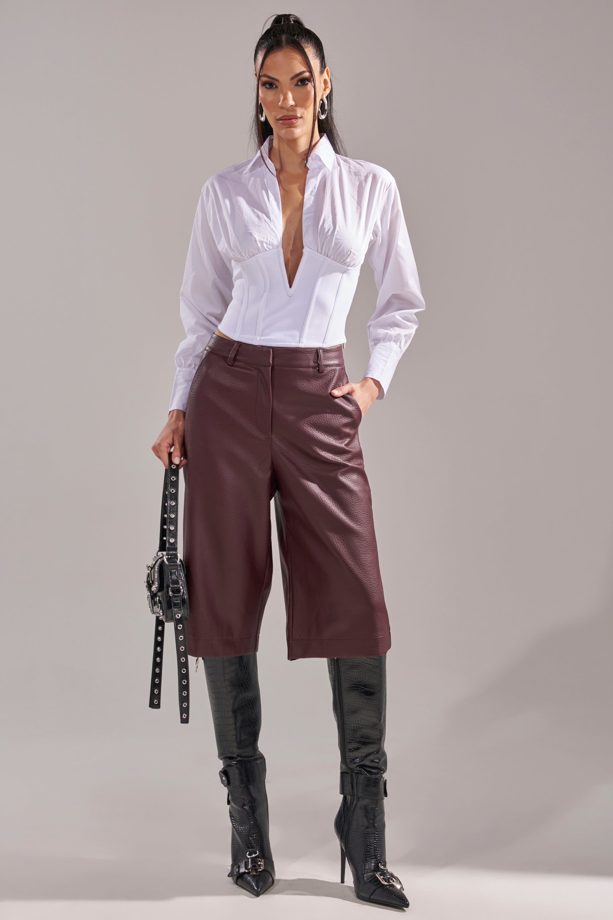 A woman stands confidently in the SNATCHED UP CORSET TOP, paired with burgundy knee-length leather shorts, black knee-high boots, and a black studded handbag. 7