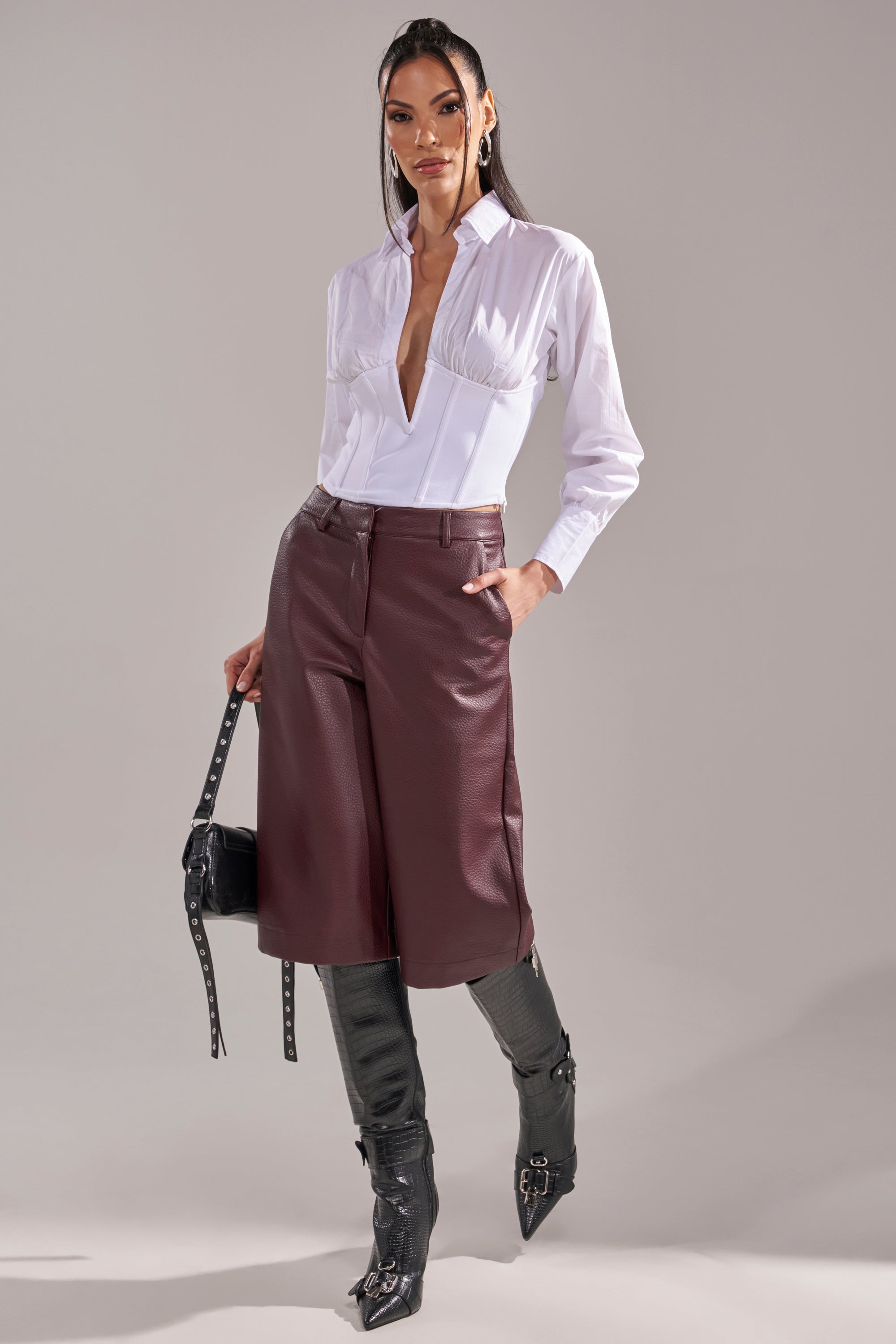 A woman stands confidently in the SNATCHED UP CORSET TOP, burgundy leather shorts, tall black boots, and holds a black studded handbag with one hand in her pocket against a plain background. 8