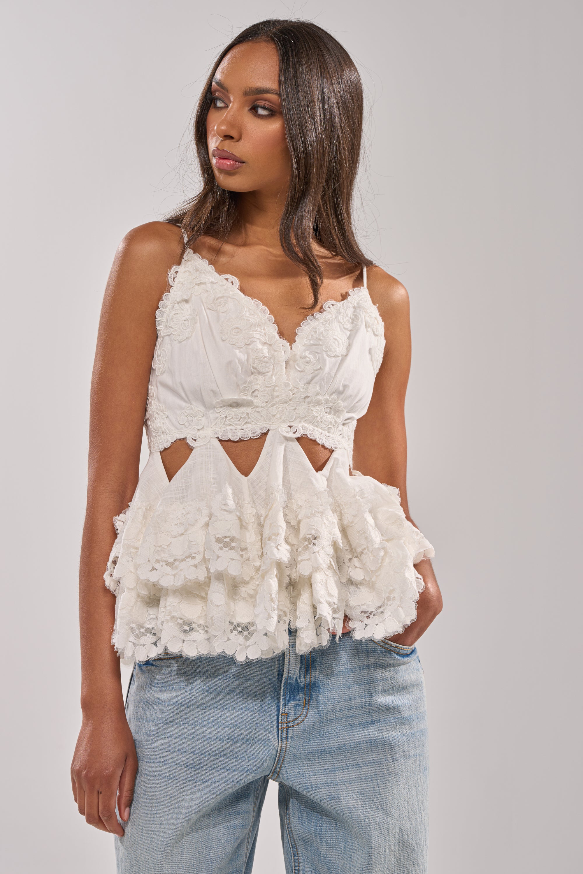 A woman models the LOTS OF LOVE TO GIVE TO YOU LACE BLOUSE, a sleeveless white floral lace top with cut-out details and a ruffled peplum hem, styled with light blue jeans against a plain light gray background. 1