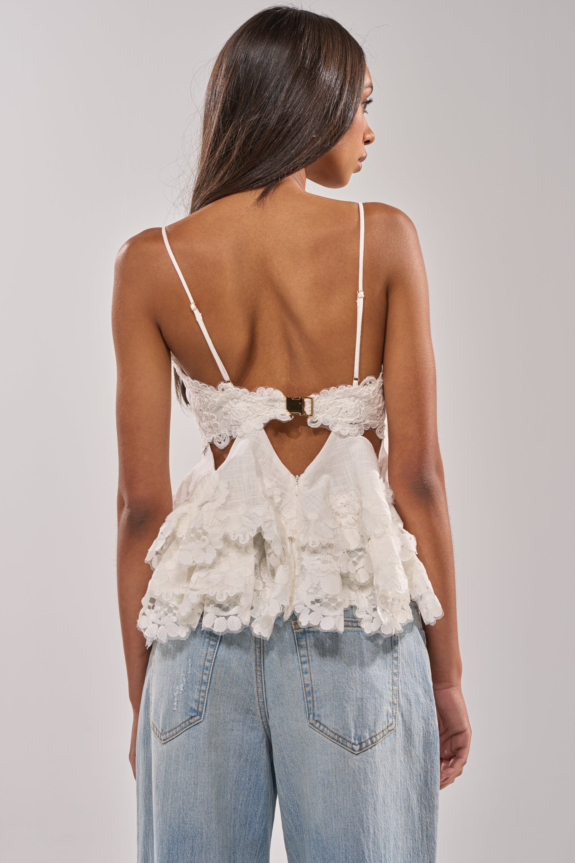 A woman with long, straight hair is seen from the back wearing the LOTS OF LOVE TO GIVE TO YOU LACE BLOUSE, featuring a floral lace peplum hem, thin straps, cutout detail with back clasp, paired with light blue high-waisted jeans. 3