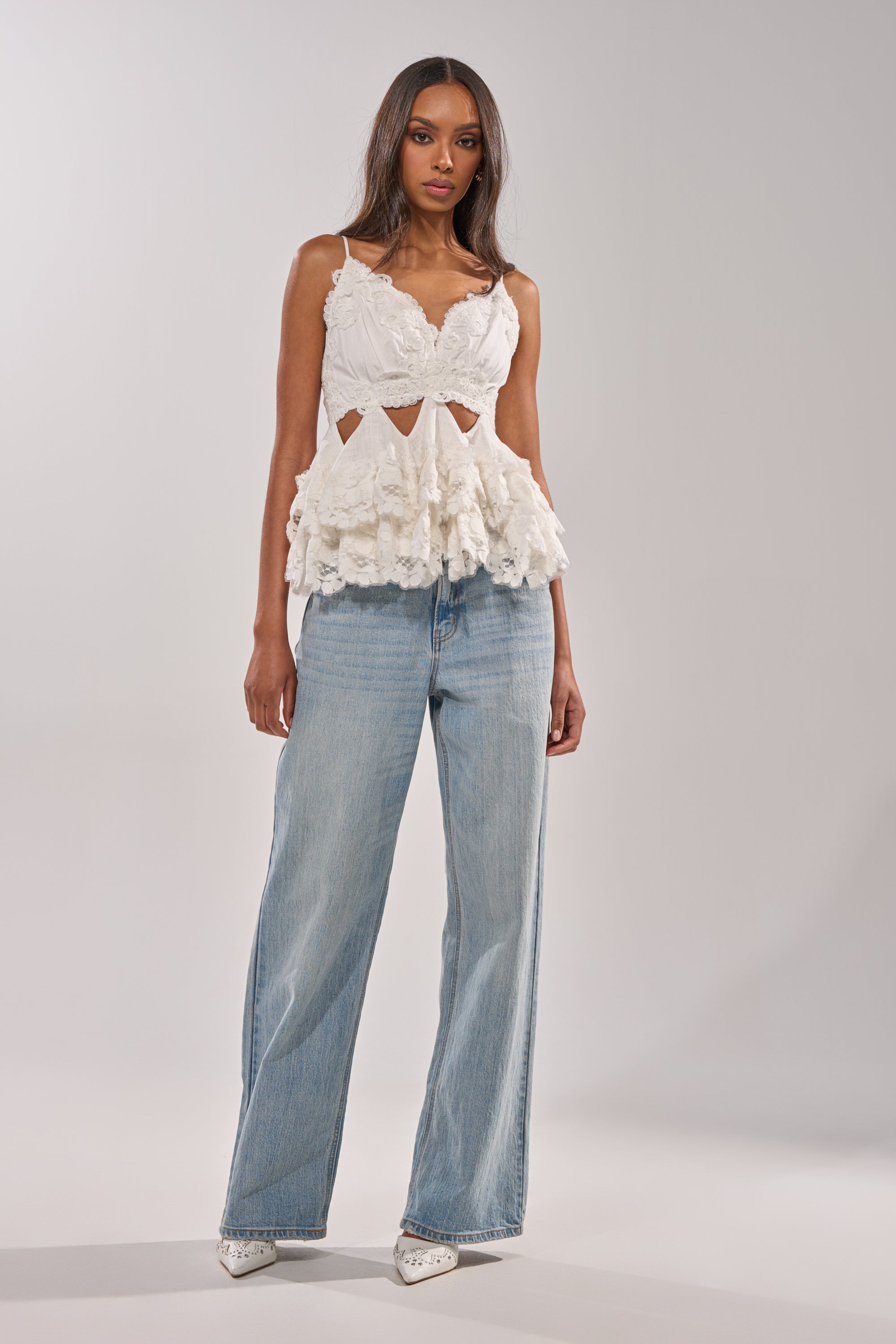 A woman stands against a neutral background wearing the LOTS OF LOVE TO GIVE TO YOU LACE BLOUSE—white, ruffled, sleeveless, with cut-out details—paired with light blue wide-leg jeans and white pointed-toe pumps. 4