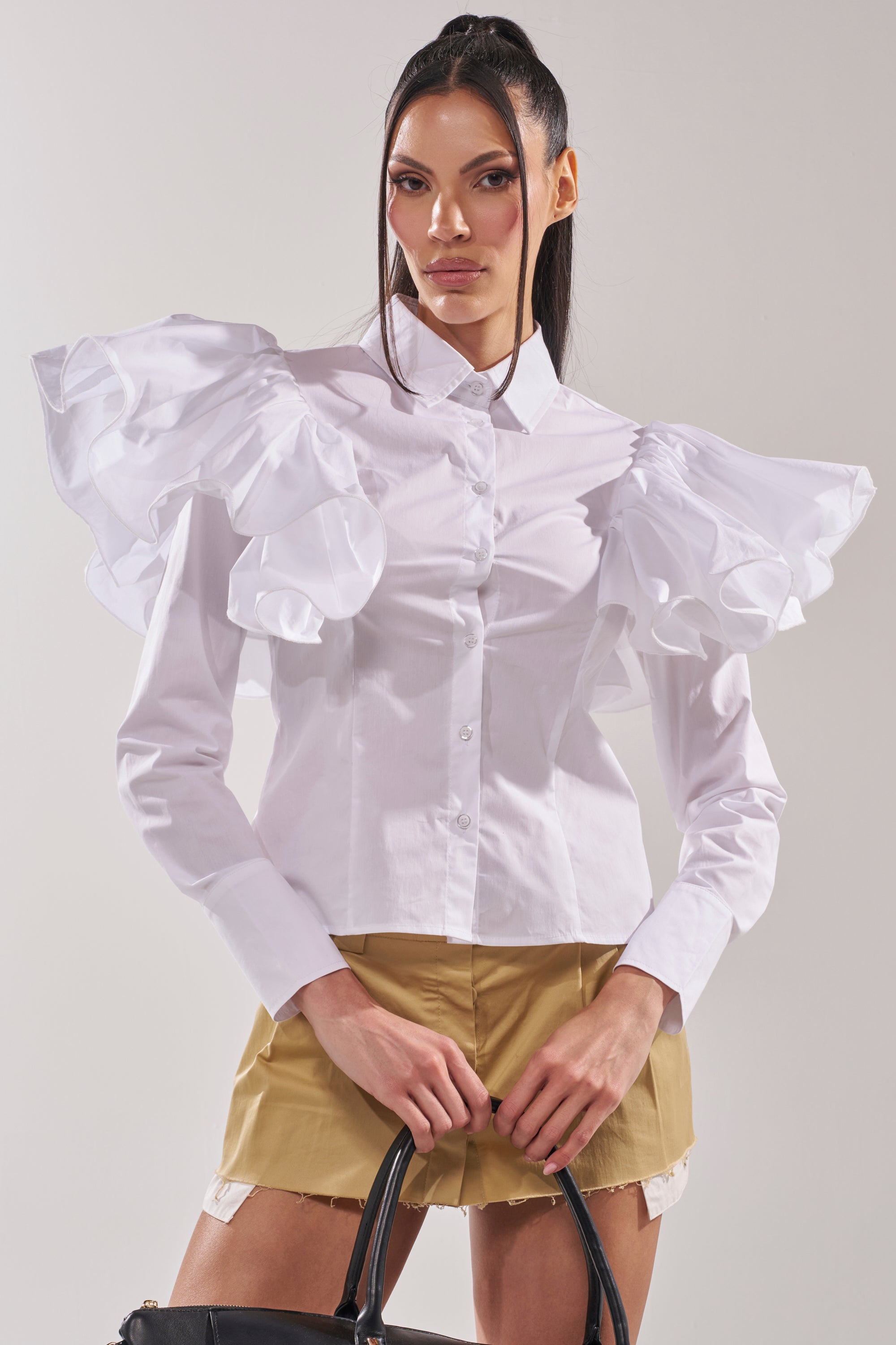 A woman wears the NO BIG DEAL BUTTON DOWN, a white blouse with ruffle shoulders, paired with tan shorts and a black handbag, posing confidently against a light background for an elevated modern workwear look. 0