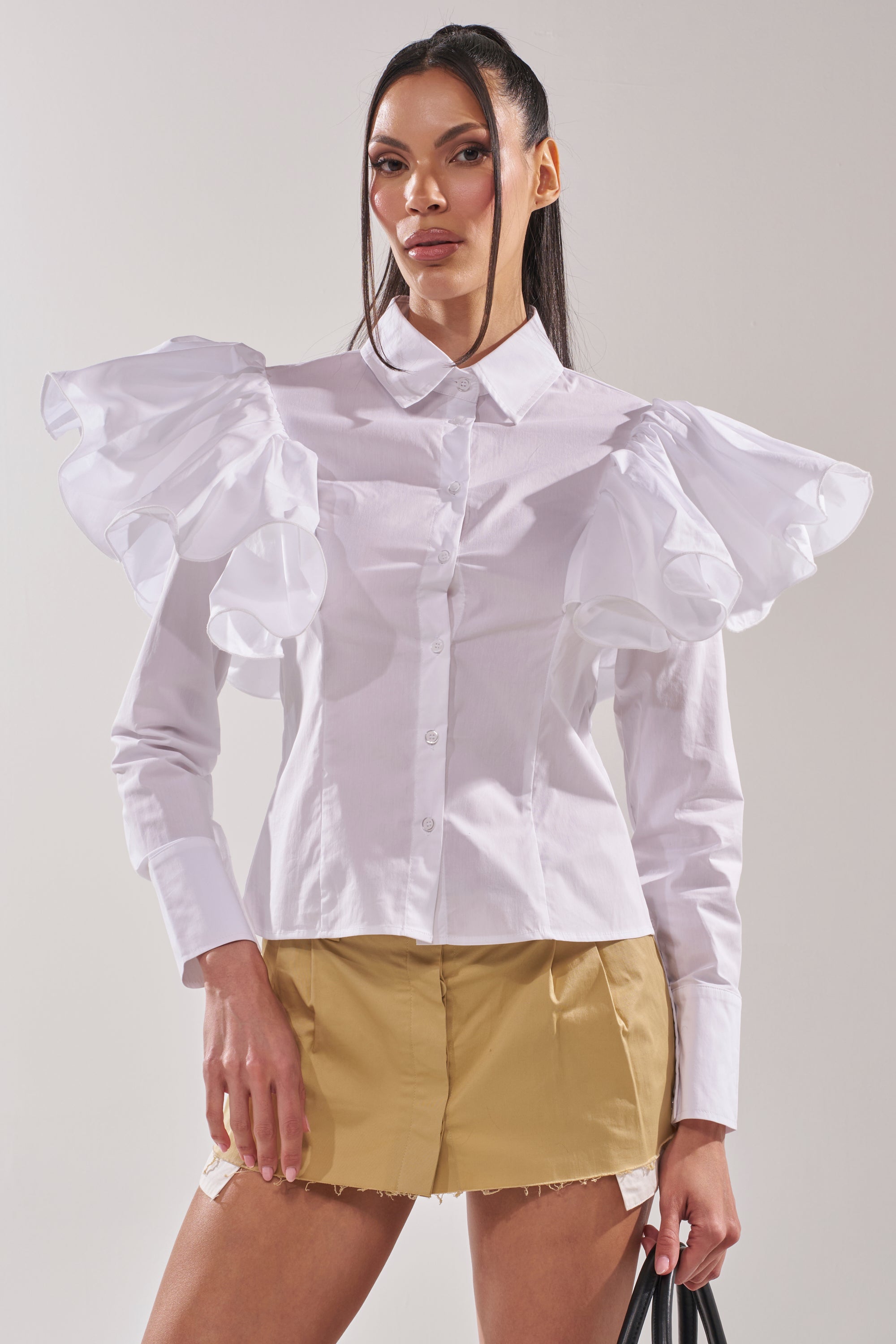 A woman poses against a plain light background wearing the NO BIG DEAL BUTTON DOWN, a white ruffle shoulder blouse with exaggerated sleeves, paired with a tan raw hem mini skirt and holding a black bag. 1