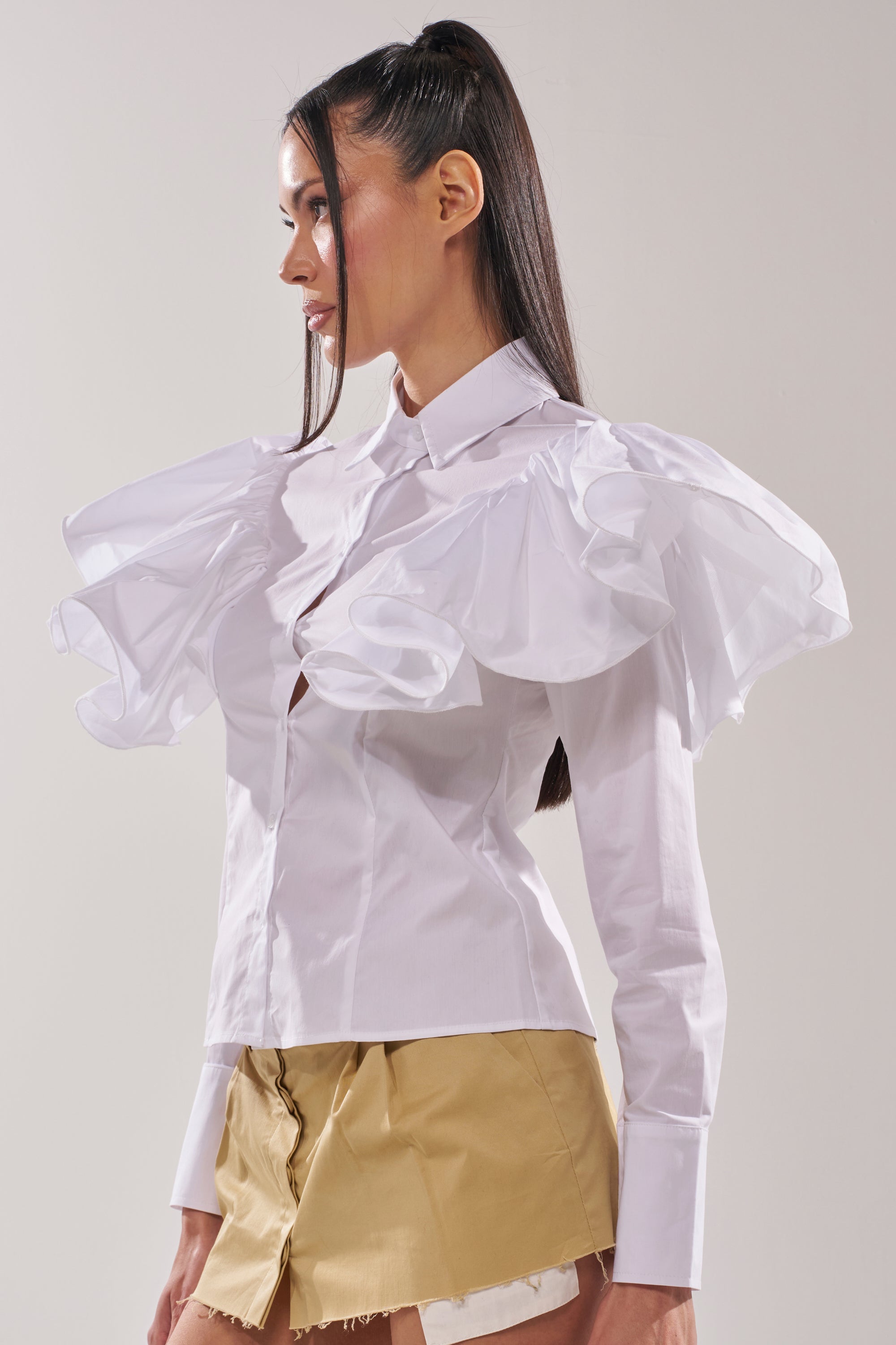 A woman with long straight hair stands in profile against a plain background, wearing the NO BIG DEAL BUTTON DOWN blouse with ruffle shoulders, puffed sleeves, and a beige mini skirt featuring a layered asymmetrical hem. 2