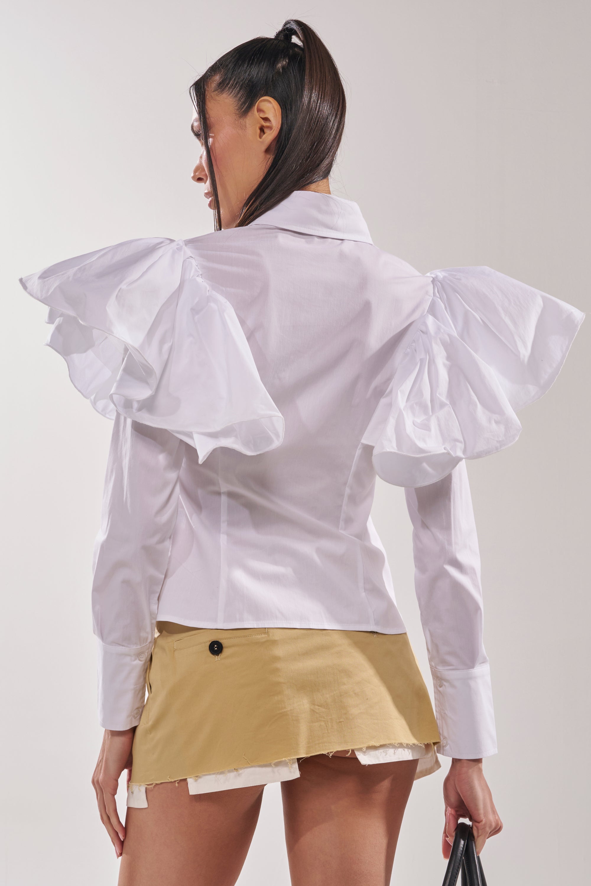 A woman with a high ponytail, facing away, wears the NO BIG DEAL BUTTON DOWN blouse with ruffle shoulders and puff sleeves, paired with a beige frayed mini skirt. She holds a black item in her right hand. 3