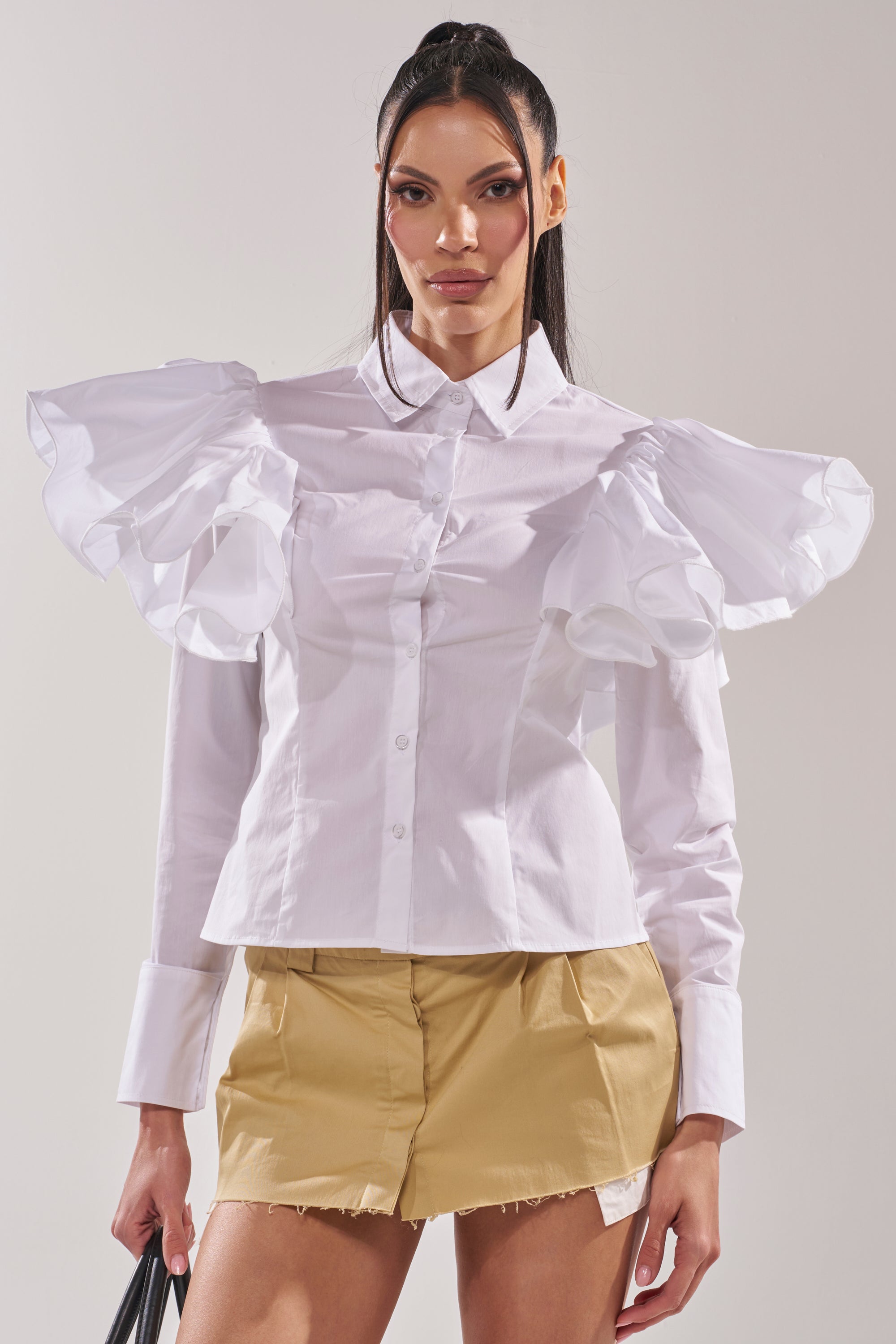 A woman wears the NO BIG DEAL BUTTON DOWN, a white blouse with ruffle shoulders, paired with tan shorts and a black handbag—a chic update on modern workwear. 4