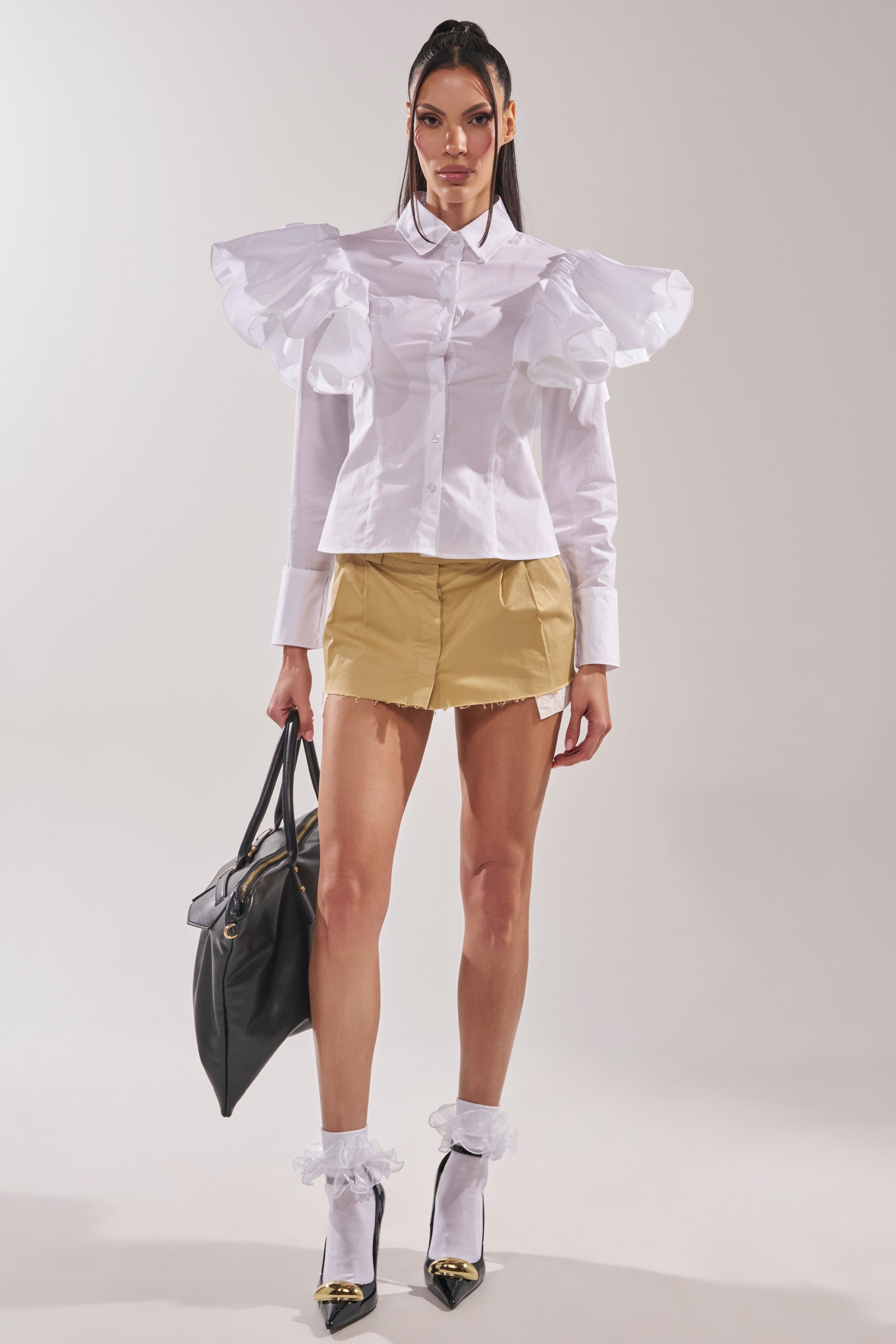 A woman stands against a plain background wearing the NO BIG DEAL BUTTON DOWN, a white blouse with large ruffled sleeves, paired with a short tan skirt, sheer socks with white ruffles, black heels, and carrying a large black handbag. 7