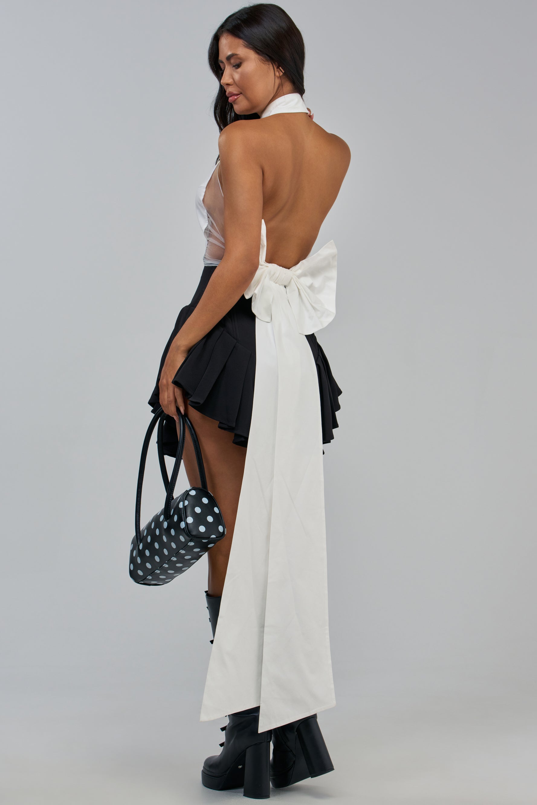 A woman wearing the GIVE IT UP POPLIN TOP—a white crop top with an open back and large white bow—paired with a black pleated mini skirt, knee-high boots, and carrying a black and white polka dot handbag stands with her back partially turned.