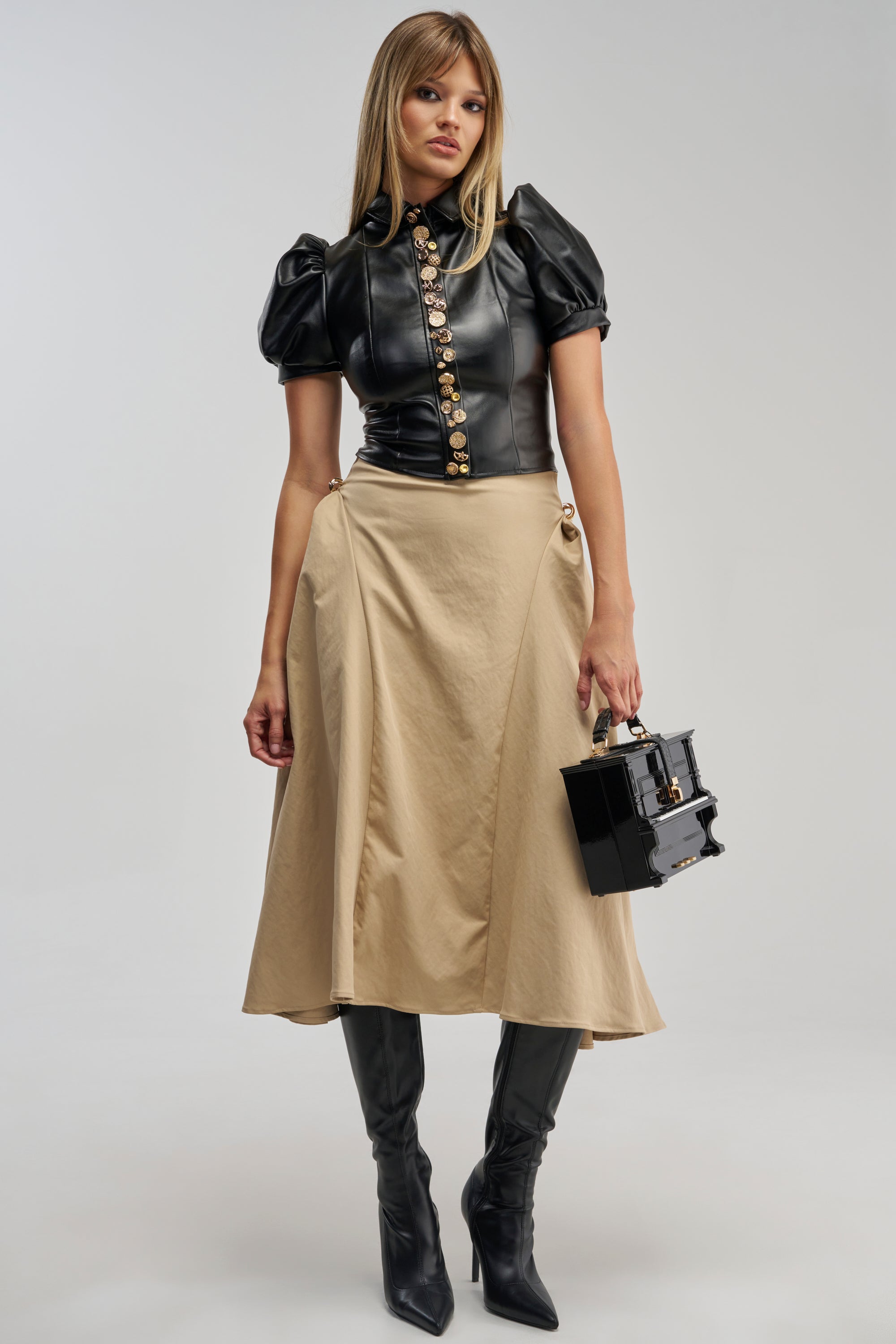 A woman stands against a plain light background wearing the IT’S MY TIME MAXI SKIRT in beige, paired with a short-sleeve black leather top featuring gold buttons, black knee-high boots, and holding a small black handbag.