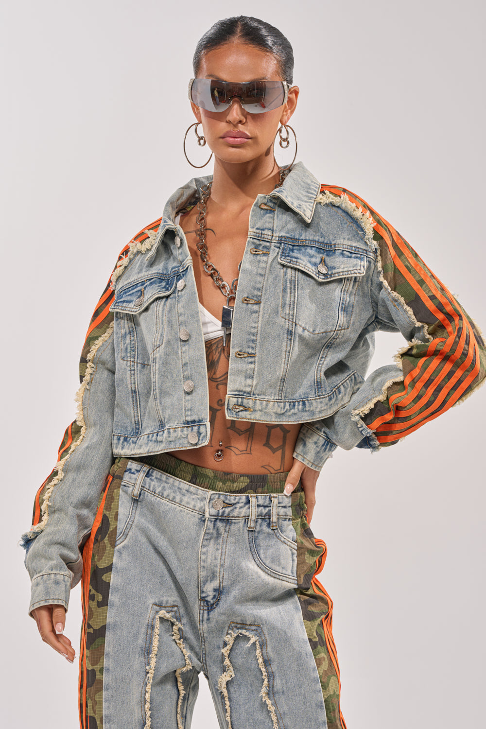 A woman in sunglasses, large hoop earrings, and a bold streetwear outfit featuring the ALL AROUND THE WORLD AGAIN JACKET IN LIGHT BLUE DENIM stands confidently against a plain background. 0