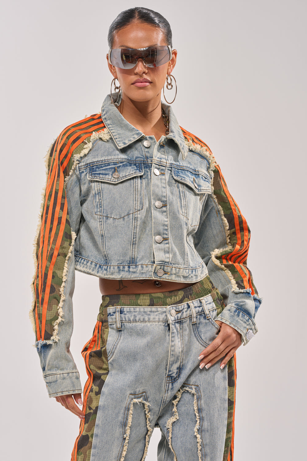 A person shows off street style in sunglasses, hoop earrings, the ALL AROUND THE WORLD AGAIN JACKET IN LIGHT BLUE DENIM with orange stripes and frayed edges, plus matching jeans with bold orange stripes and camo accents. 3
