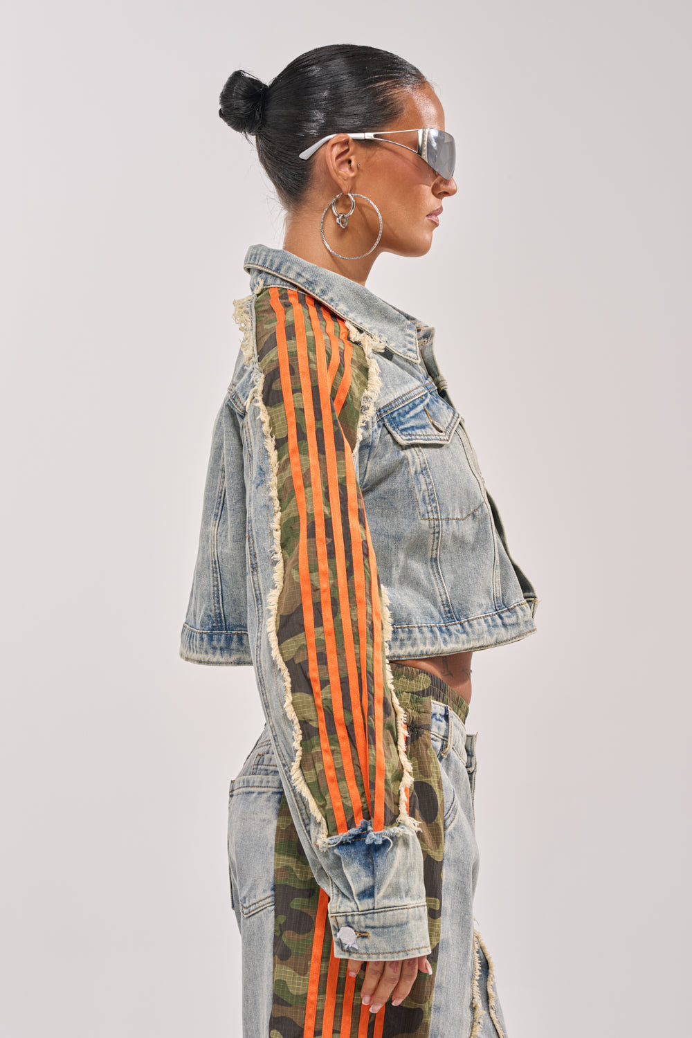 A woman in profile wears oversized sunglasses, large hoop earrings, and the ALL AROUND THE WORLD AGAIN JACKET IN LIGHT BLUE DENIM with orange stripes, paired with camo accent pants for a bold streetwear look. Her hair is slicked back in a bun. 4
