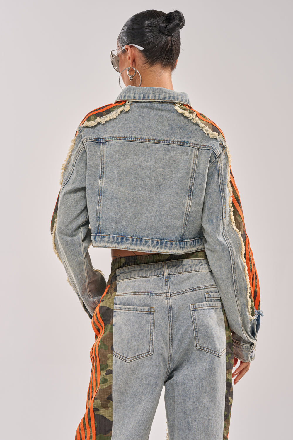 Wearing the ALL AROUND THE WORLD AGAIN JACKET IN LIGHT BLUE DENIM, styled with high-waisted jeans with camo accents and orange stripes, a person with hair in a bun poses back to the camera in bold streetwear. 5