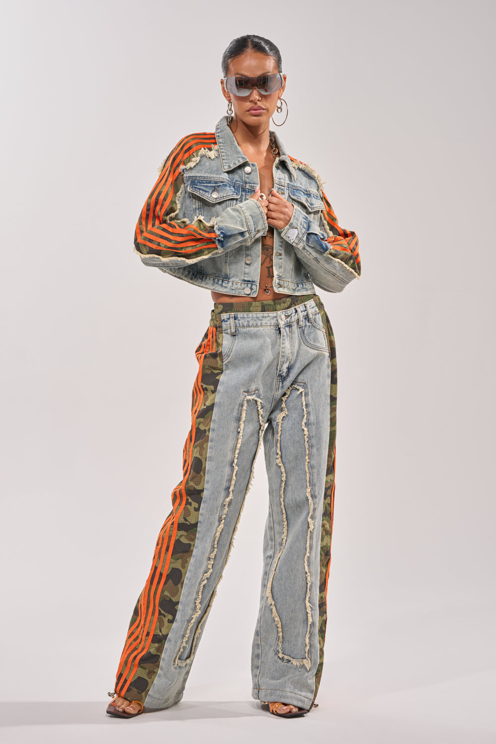 A person wearing sunglasses poses in the ALL AROUND THE WORLD AGAIN JACKET IN LIGHT BLUE DENIM, styled with distressed jeans featuring orange stripes, camo accents, a crop top, and open-toe shoes against a plain background. 6