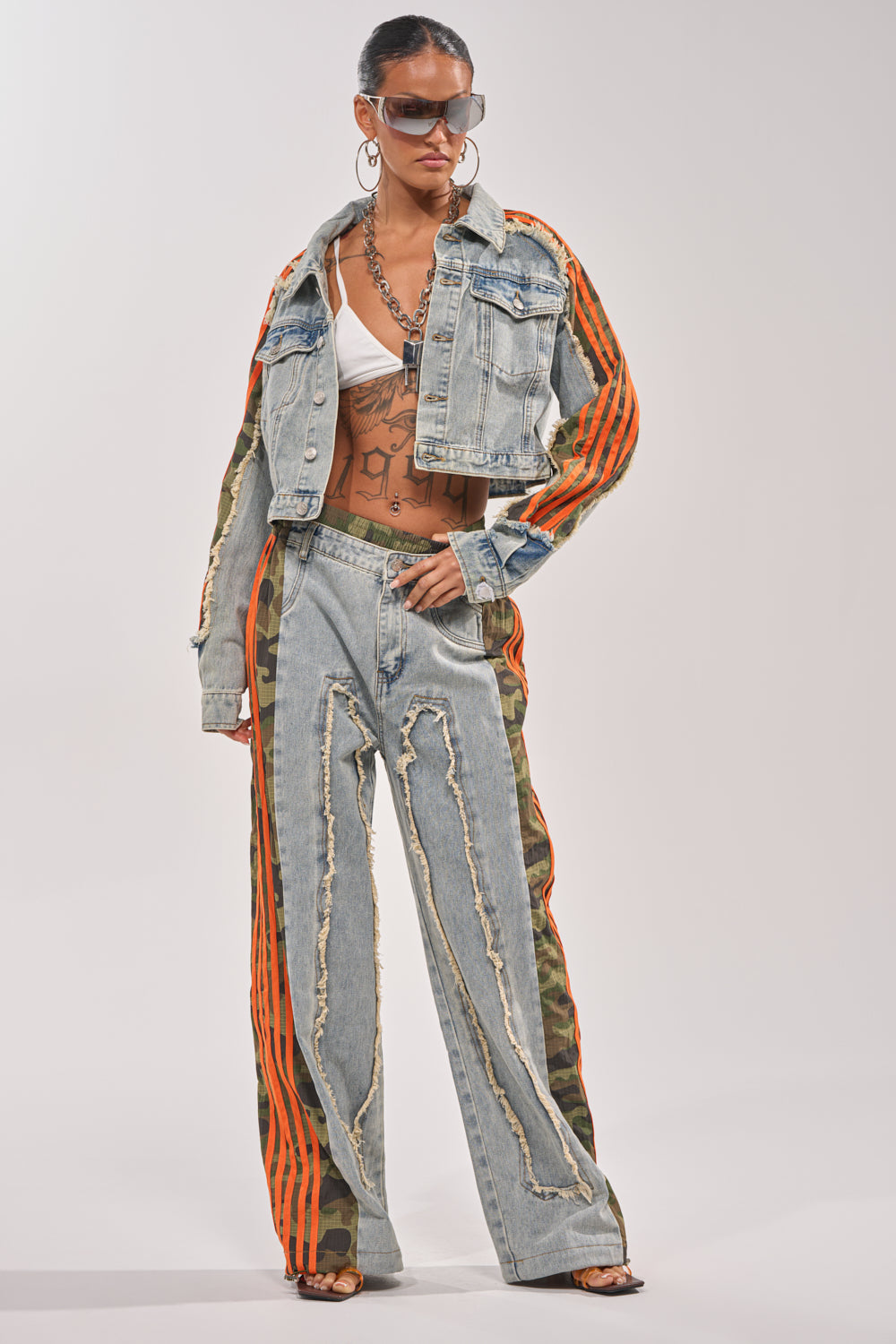 A person in sunglasses and the ALL AROUND THE WORLD AGAIN JACKET IN LIGHT BLUE DENIM with bold orange stripes, styled with a white bralette, distressed denim pants featuring orange stripes and camo accents, hoop earrings, and layered necklaces exudes effortless streetwear cool. 7