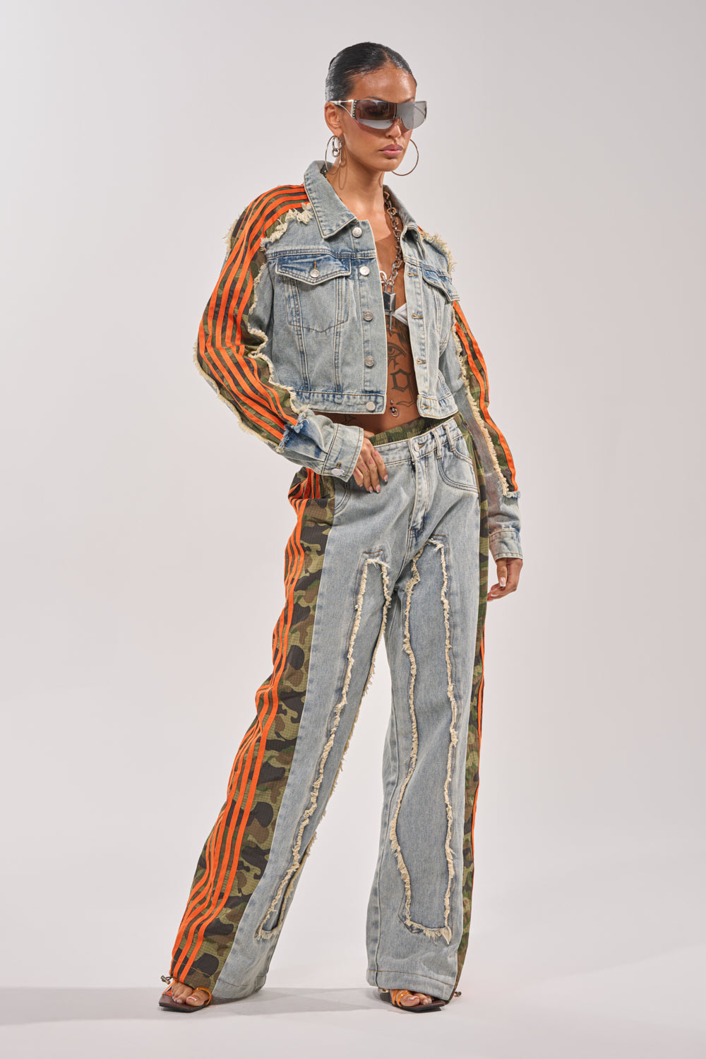 A person poses confidently in the ALL AROUND THE WORLD AGAIN JACKET IN LIGHT BLUE DENIM, paired with jeans featuring orange stripes and camo side panels, sunglasses, hoop earrings, and heeled sandals against a plain background. 8