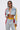 A woman exudes street-style confidence in sunglasses, a white bralette, and the ALL AROUND THE WORLD AGAIN DENIM JACKET with red and yellow striped sleeves, paired with matching high-waisted distressed jeans against a plain light background.