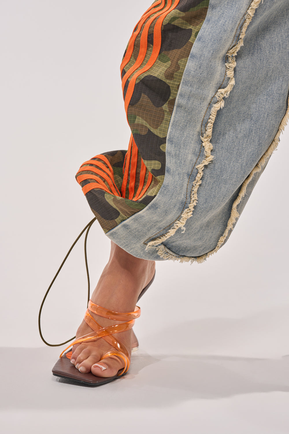 A close-up of a foot in an orange strappy sandal with a square toe, wearing ALL AROUND THE WORLD AGAIN RELAXED FIT DENIM—wide-leg camo denim pants with frayed hems and orange striped accents. The background is plain and light-colored. 9