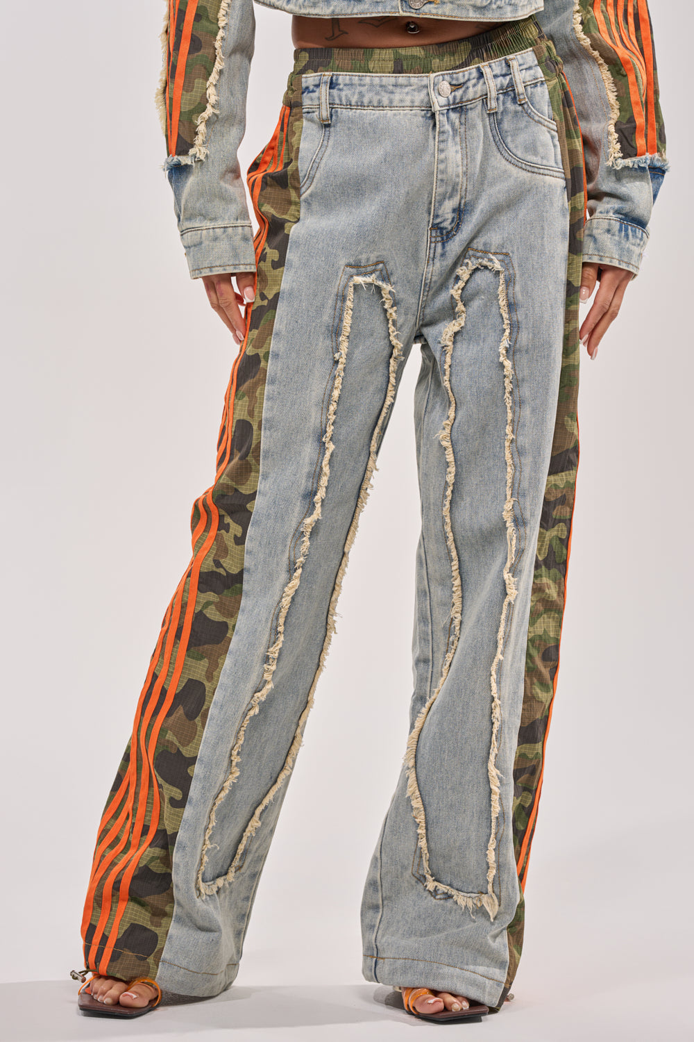 Someone wearing the ALL AROUND THE WORLD AGAIN RELAXED FIT DENIM, featuring distressed patchwork with light blue fronts, camo denim side panels, bright orange stripes, frayed edges, and open-toed heels. 1