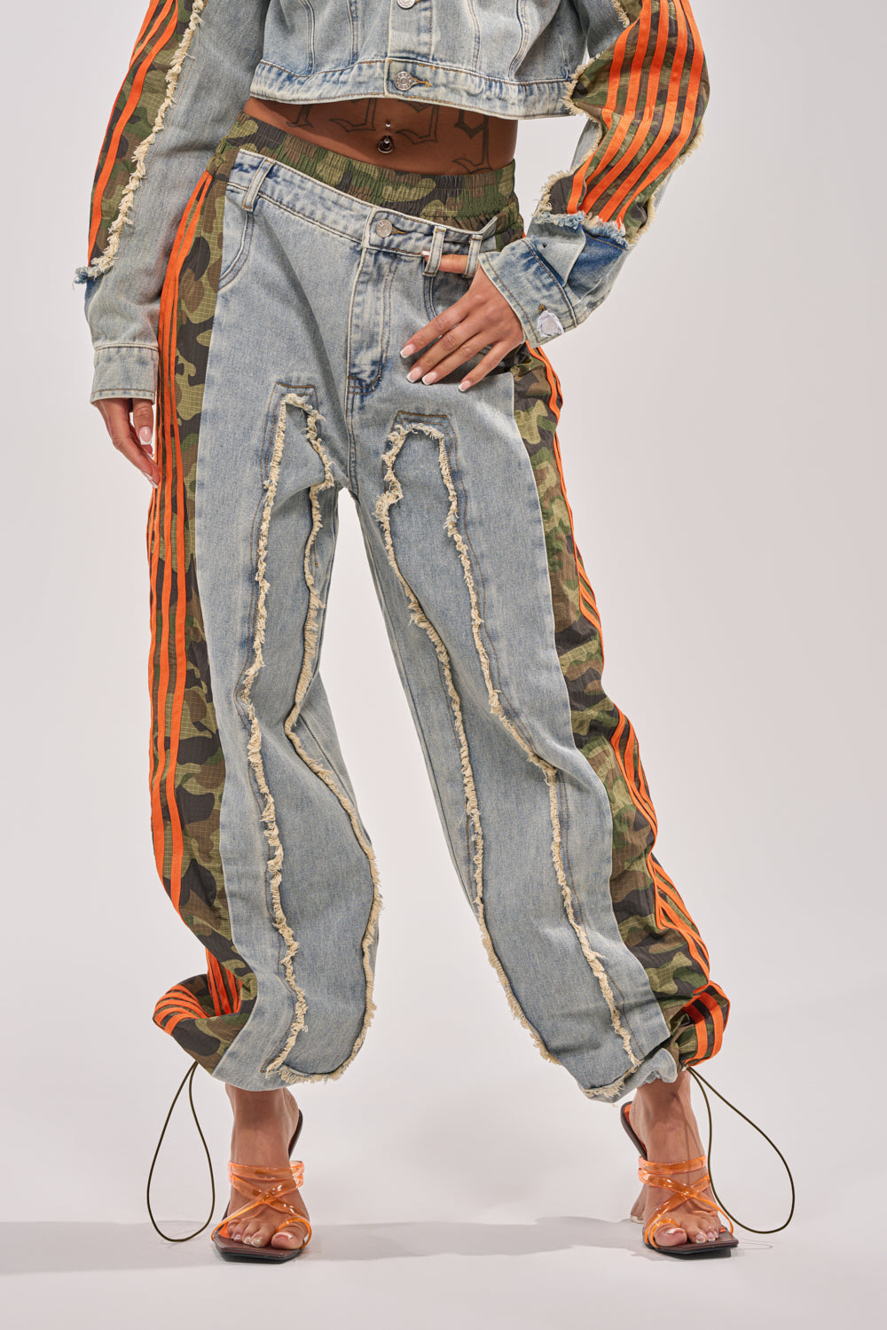 Wearing the ALL AROUND THE WORLD AGAIN RELAXED FIT DENIM with orange stripes and camo panels, paired with a cropped denim jacket and orange strappy heels, this look exudes bold streetwear style against a simple backdrop. 2