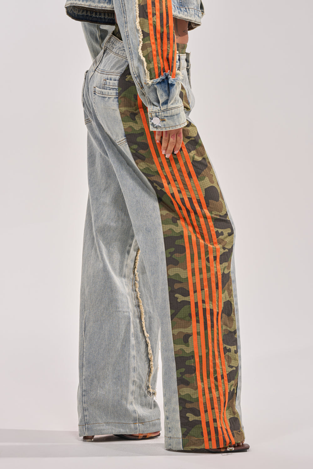 Model showcases the ALL AROUND THE WORLD AGAIN RELAXED FIT DENIM—camo wide-leg pants with denim and camouflage patchwork, bold orange seam stripes, and distressed hems. Only the lower body appears against a plain, light background. 4