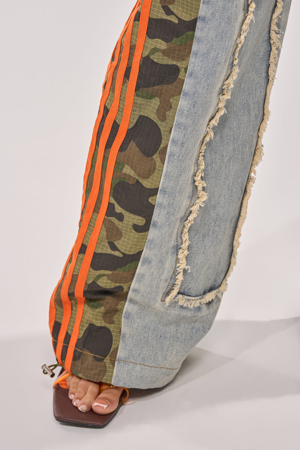 A close-up of the ALL AROUND THE WORLD AGAIN RELAXED FIT DENIM with camo patchwork, orange stripes, and frayed light blue panels, styled with open-toe sandals for a bold streetwear look. 8