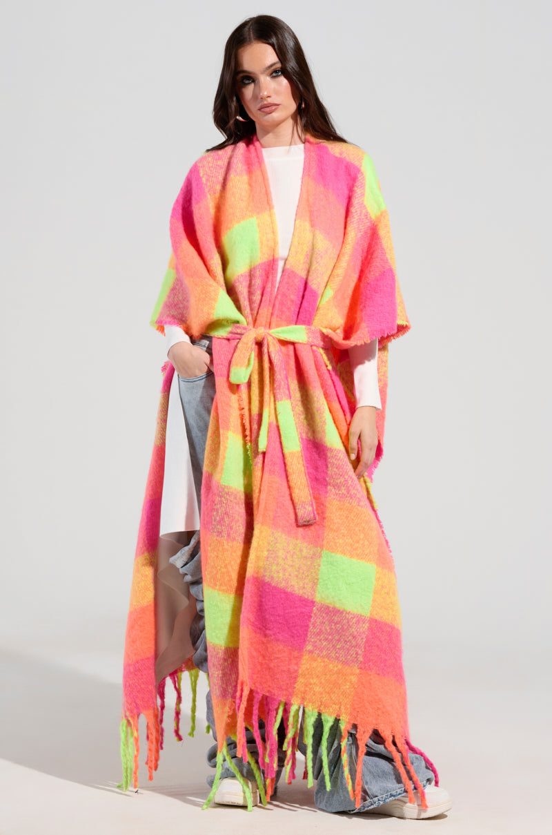A woman confidently wears the UNDER THE SUN PLAID PONCHO, showcasing its vibrant pink, orange, and green plaid design. The poncho includes fringe detailing and a belt, elegantly covering her white top and blue jeans. She stands against a light background, embodying a casual yet bold style with a touch of bohemian flair.