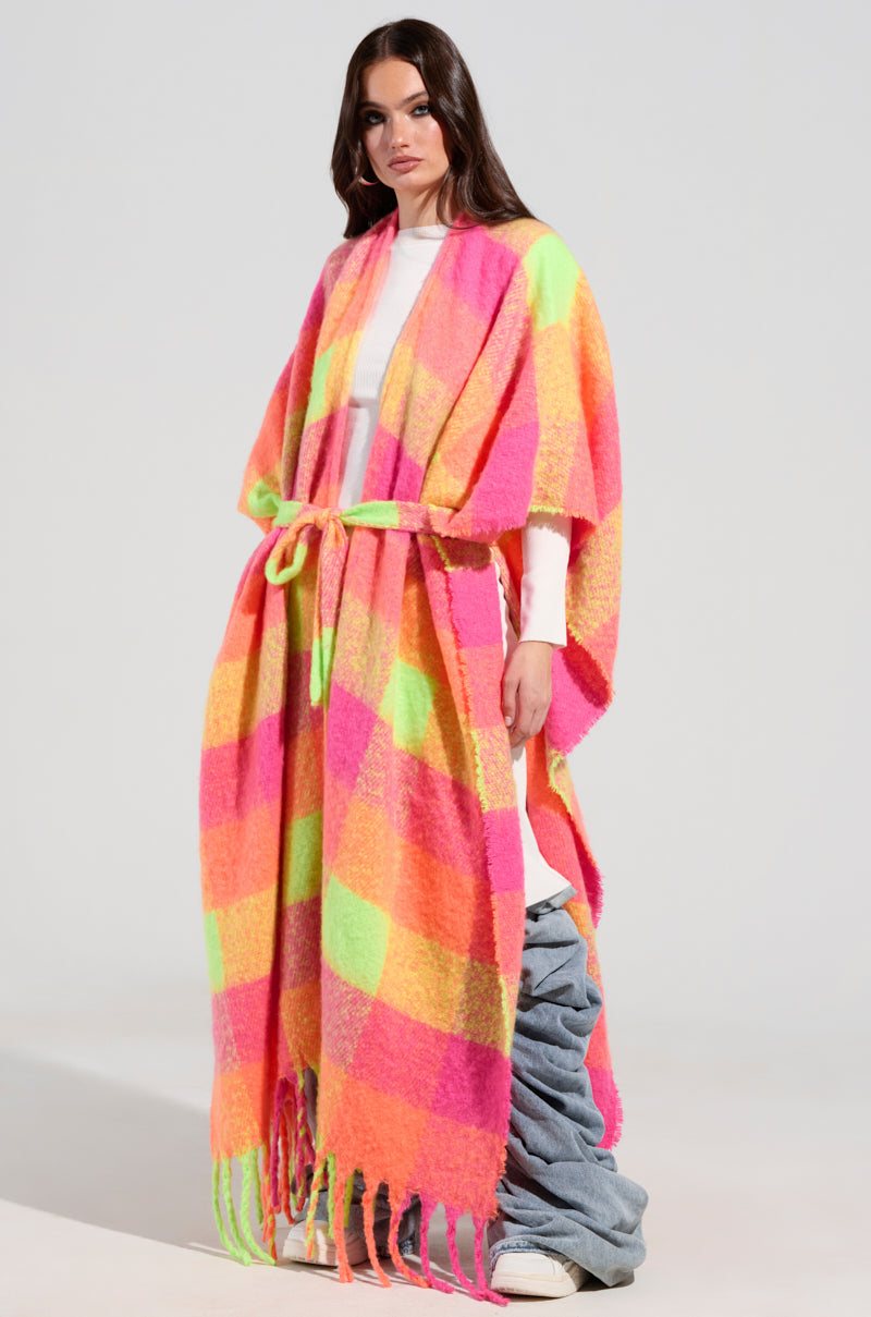 A person wearing the UNDER THE SUN PLAID PONCHO, a vibrant, multi-colored piece with fringe detailing and a belted waist over a white outfit. The poncho showcases hues of pink, orange, yellow, and green. The look is completed with light blue ruched pants and white shoes.
