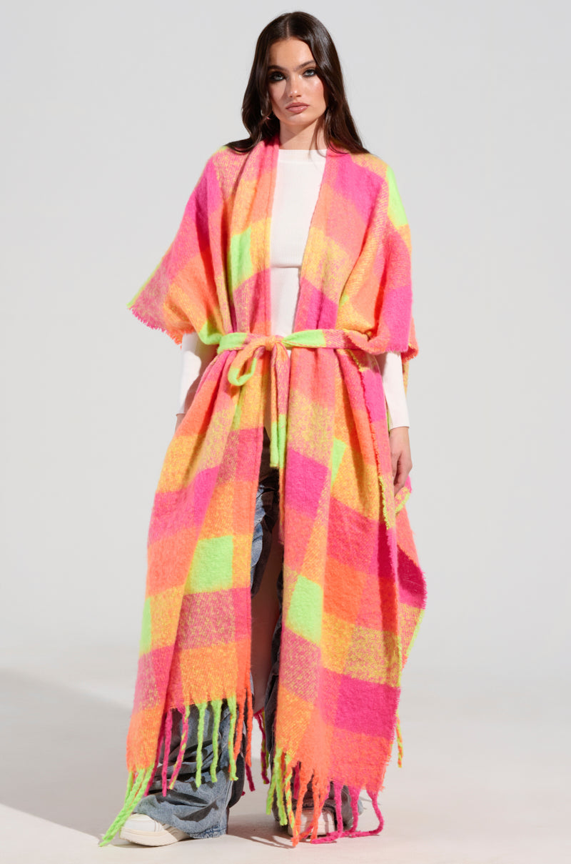 A person wears the UNDER THE SUN PLAID PONCHO, showcasing vibrant neon pink, orange, and yellow patterns over a white shirt and blue jeans. The poncho features a maxi silhouette with elegant fringe details at the bottom and is stylishly tied at the waist.