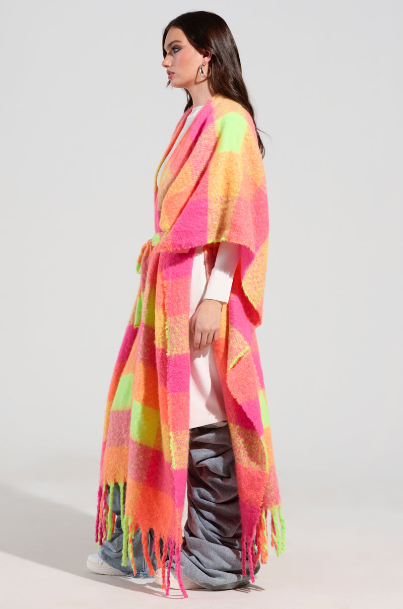 A person wearing the UNDER THE SUN PLAID PONCHO, featuring a vibrant mix of pink, orange, and green patterns, draped over a white dress and gray pants. They are standing sideways against a plain background, highlighting the playful fringe detailing of their ensemble.