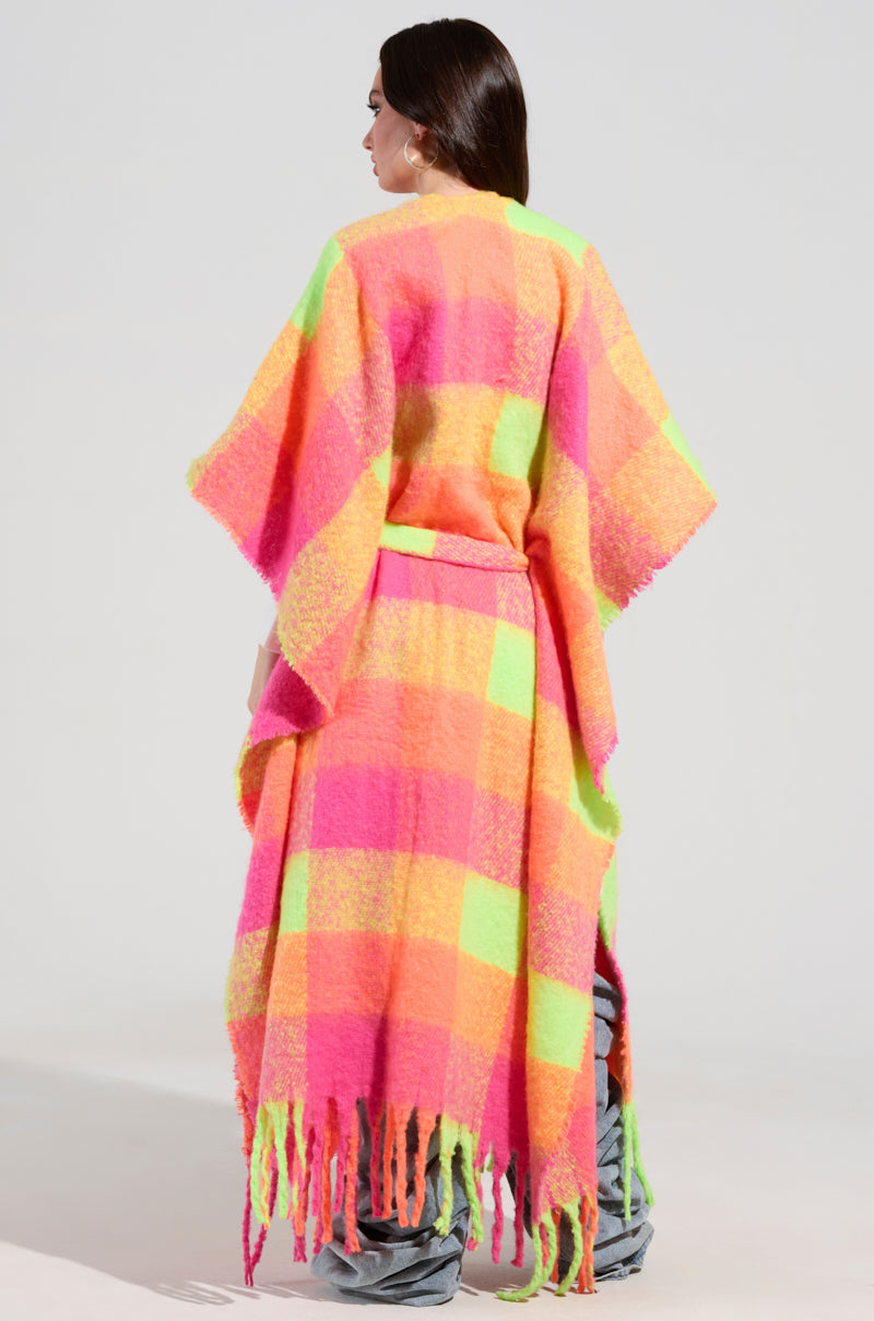 A woman models the "UNDER THE SUN PLAID PONCHO," a maxi silhouette with a long, colorful checkered pattern featuring vibrant shades of pink, orange, and yellow. The fringe detailing at the hem adds an elegant touch as she faces away from the camera to highlight the poncho's exquisite design and texture.