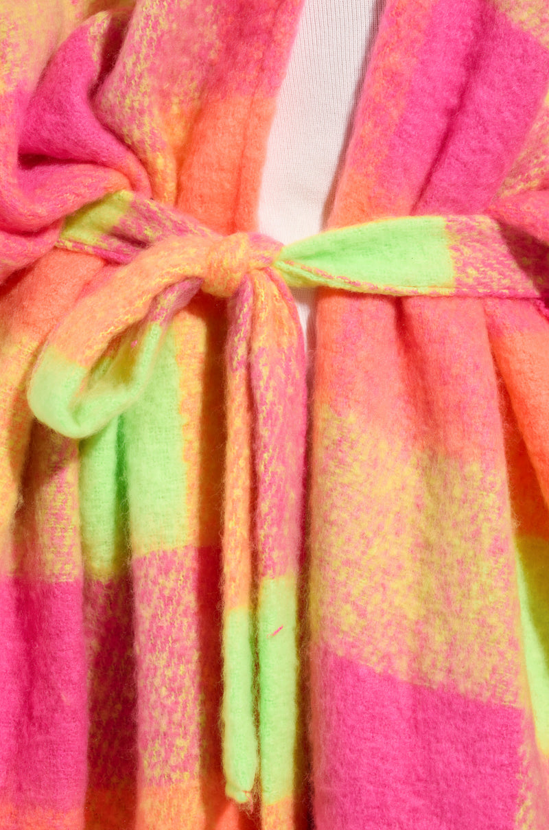 Close-up of the UNDER THE SUN PLAID PONCHO fabric tied in a bow. The material showcases a vibrant checkered pattern with shades of pink, orange, and green, creating a bright and lively appearance.