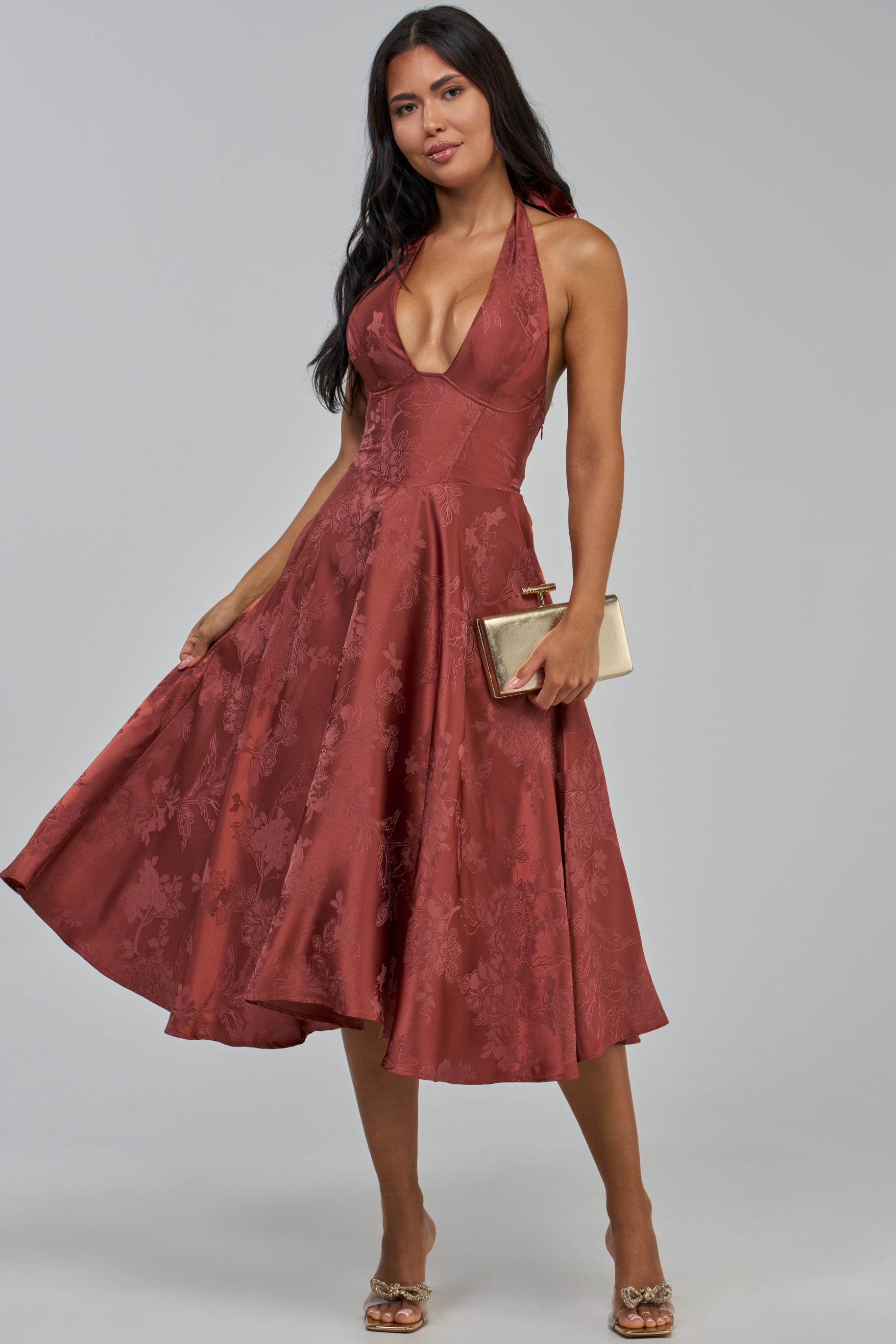 Wearing the CATCH ME LATER FLORAL MIDI DRESS—a sleeveless, deep v-neck halter style perfect as a wedding guest dress—a woman stands against a neutral background, accessorizing with a gold clutch and gold heels.
