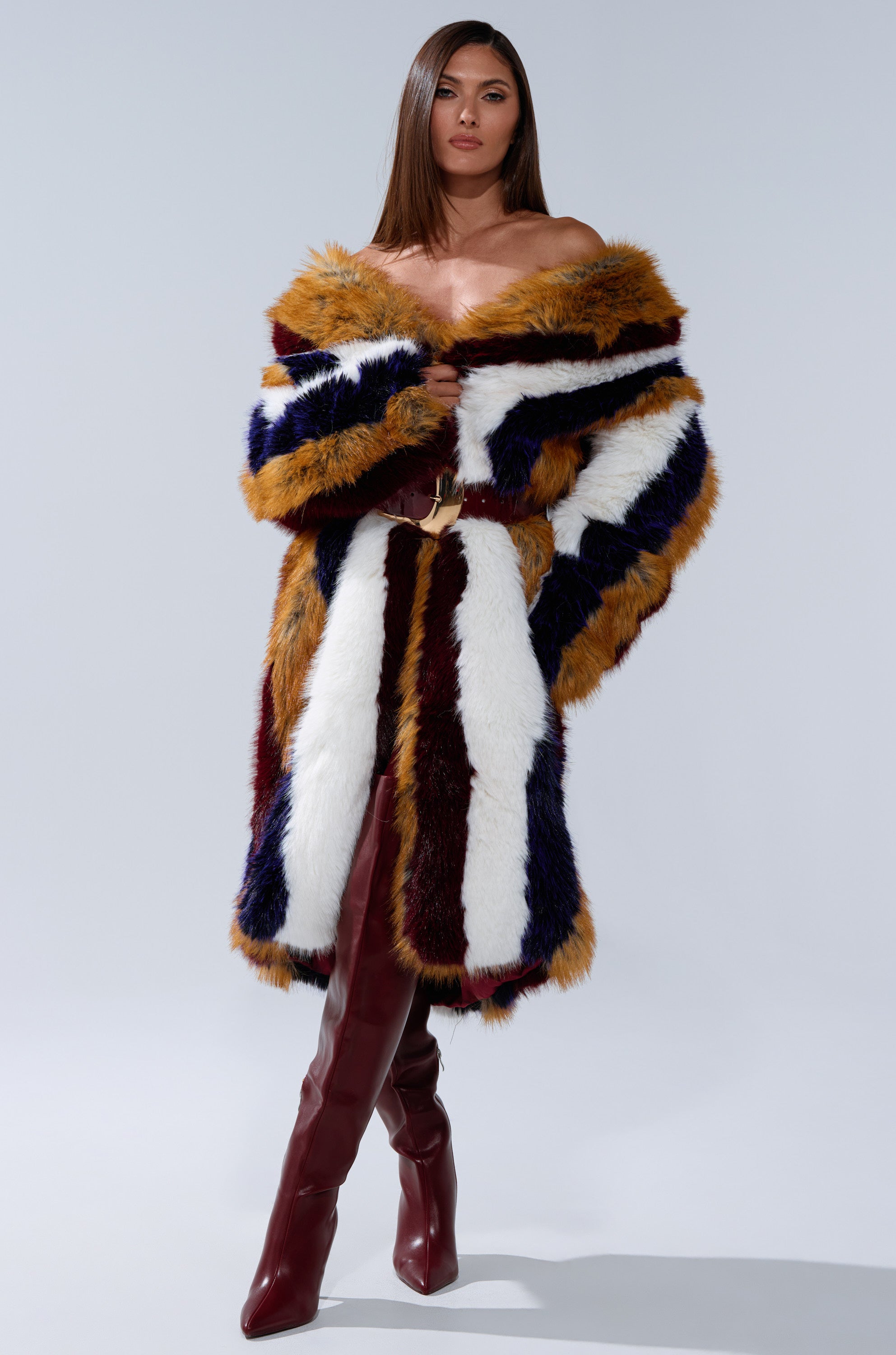 A woman models the ALAIA STRIPED FAUX FUR, an off-the-shoulder longline coat in vibrant yellow, white, navy, and burgundy stripes, paired with high burgundy boots against a simple light backdrop.