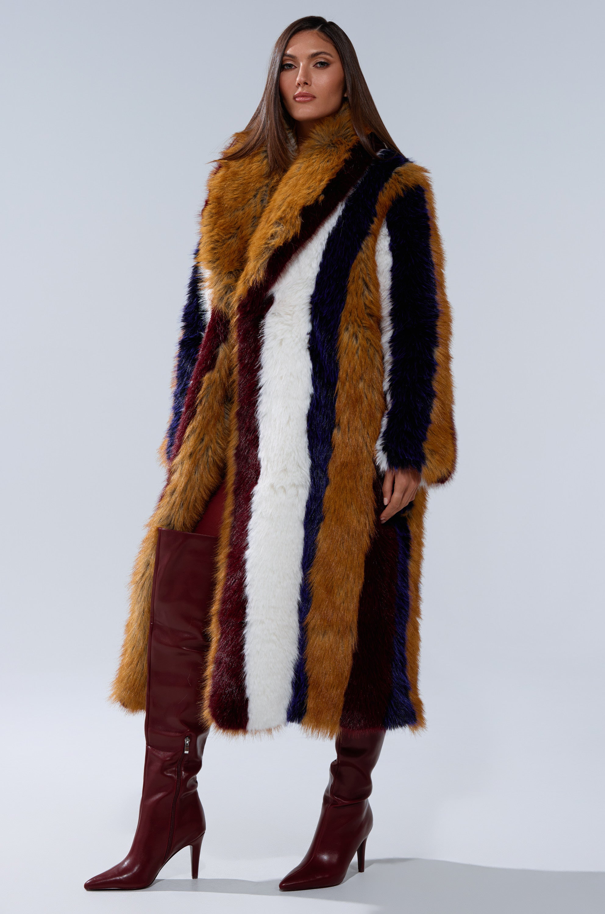A woman stands confidently in the ALAIA STRIPED FAUX FUR longline coat with mustard yellow, white, navy, and burgundy stripes, paired with matching burgundy knee-high heeled boots.