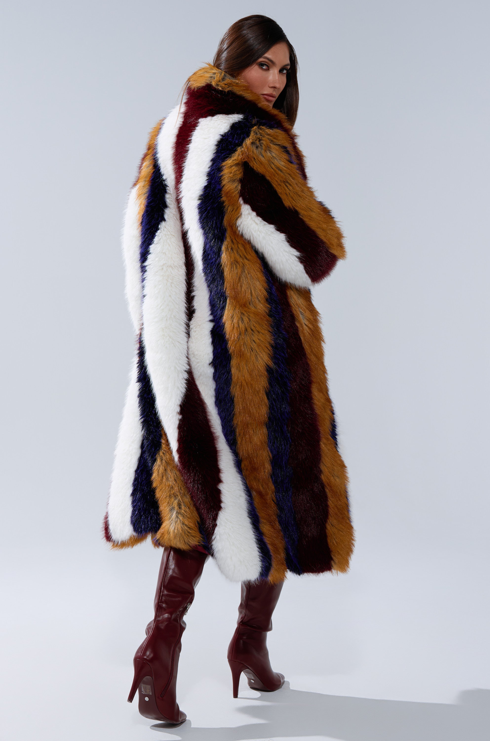 A woman poses with her back turned in the ALAIA STRIPED FAUX FUR coat featuring white, brown, navy, and burgundy stripes, paired with burgundy heeled boots against a plain background.