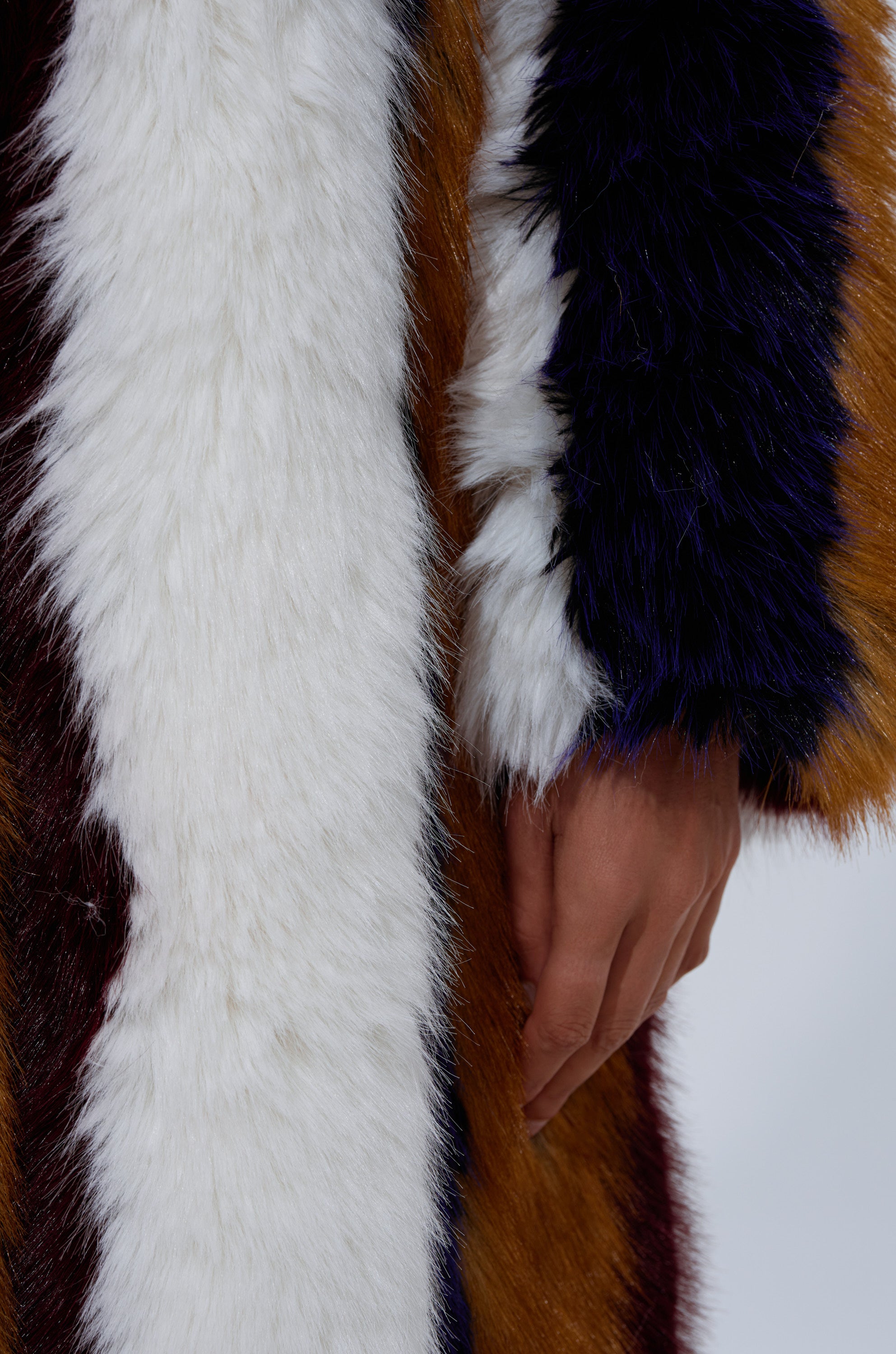 A close-up of a hand resting on the ALAIA STRIPED FAUX FUR coat, showcasing bold stripes in white, brown, black, and dark purple for a modern glam look.