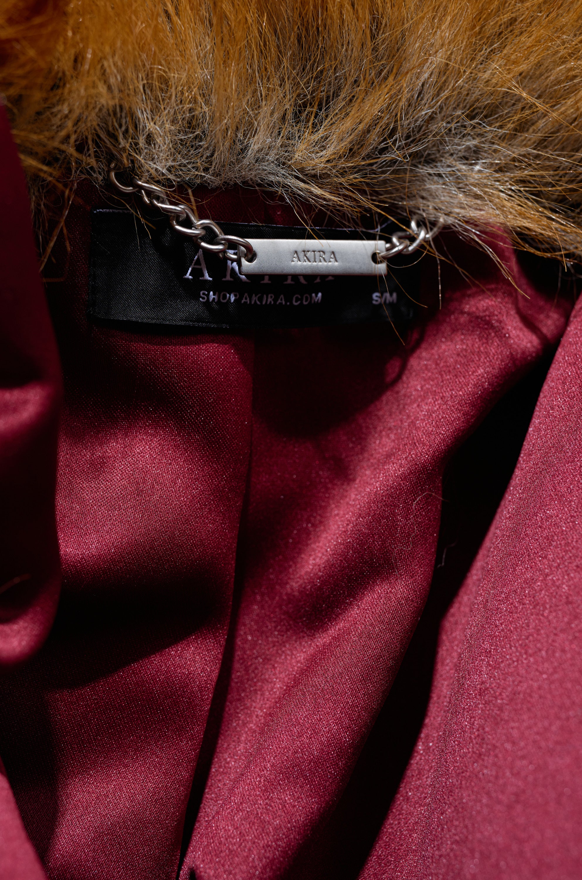 A close-up of the ALAIA STRIPED FAUX FUR coat reveals a black "AKIRA" label, fur-trimmed collar, and silver engraved chain—stylish details for those seeking modern glamor in this chic striped faux fur. "shopakira.com" appears in view.