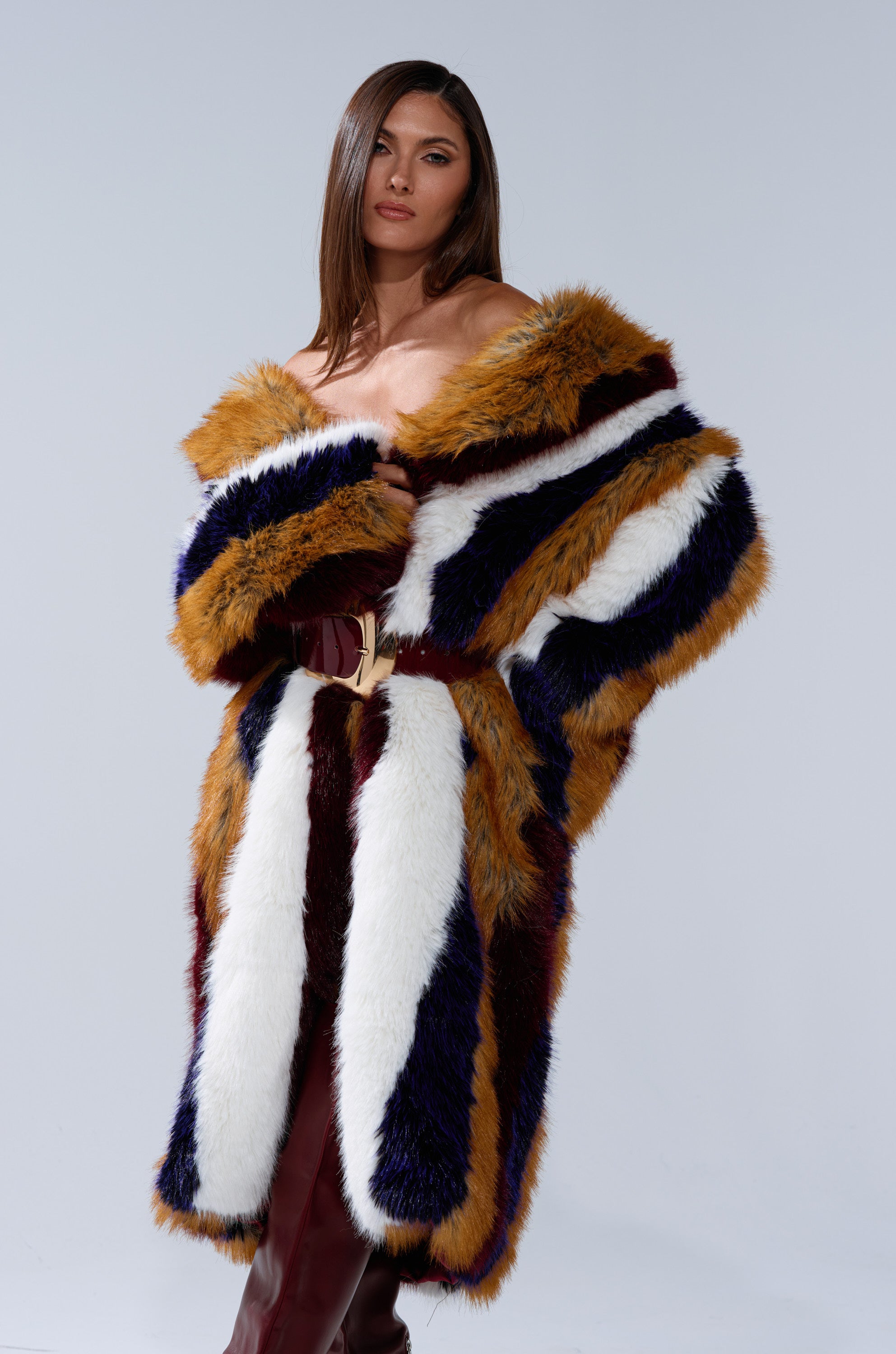 A woman with straight brown hair models the ALAIA STRIPED FAUX FUR, an off-the-shoulder coat with multicolored panels—white, brown, black, and dark blue—paired with burgundy pants against a plain light background.