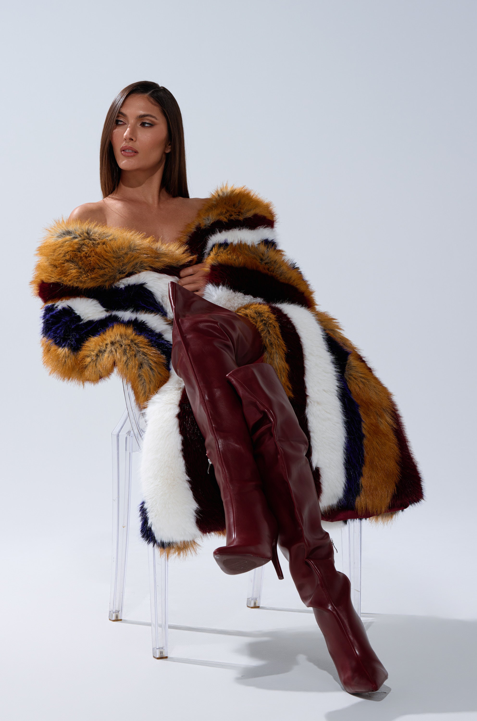 A woman sits on a clear chair in the ALAIA STRIPED FAUX FUR coat and tall, burgundy leather boots, her straight hair parted to the side as she poses confidently against a plain background.
