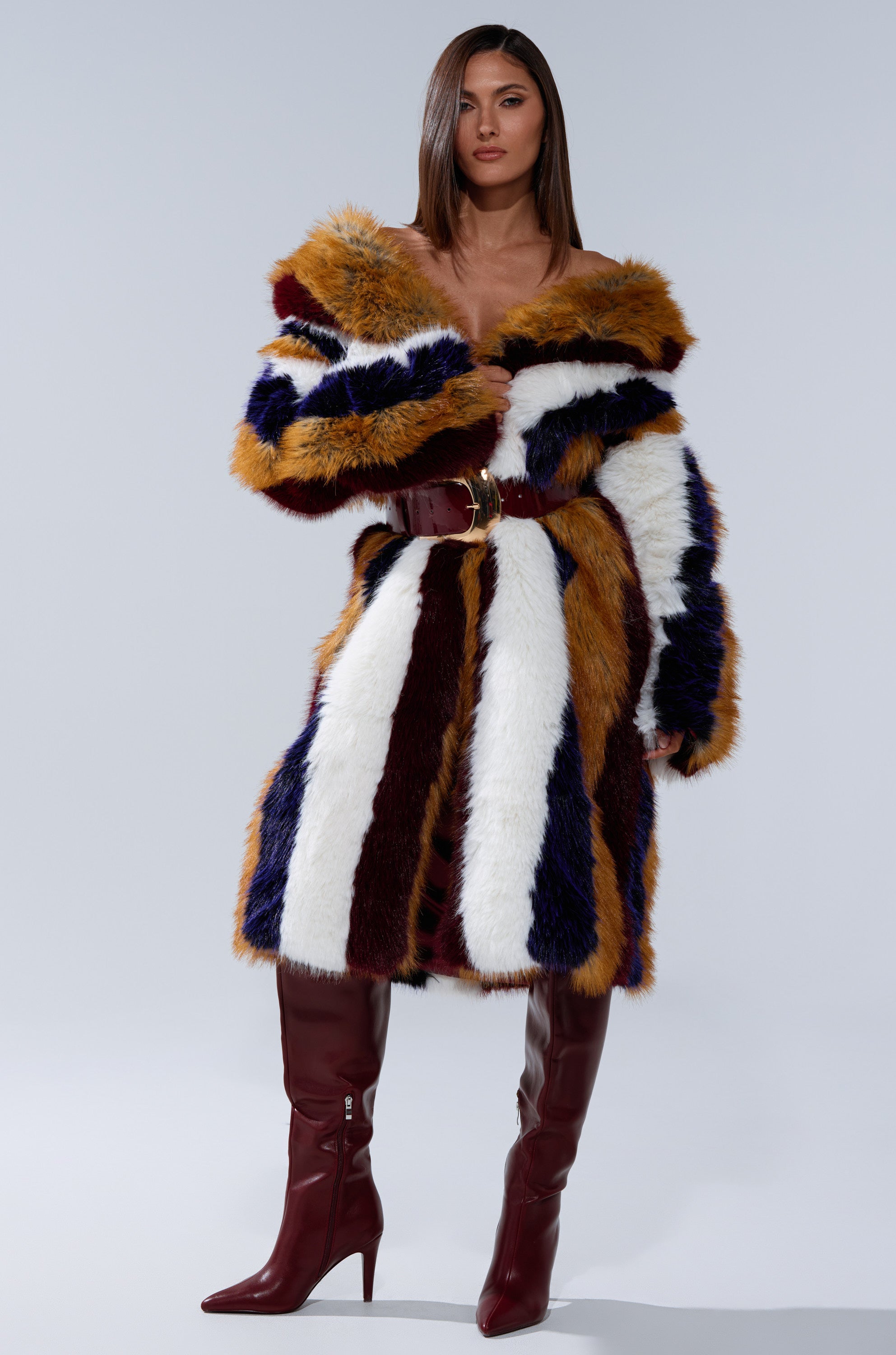 A woman with straight brown hair models the ALAIA STRIPED FAUX FUR coat in brown, white, blue, and burgundy, styled with knee-high burgundy boots, standing against a plain gray backdrop.
