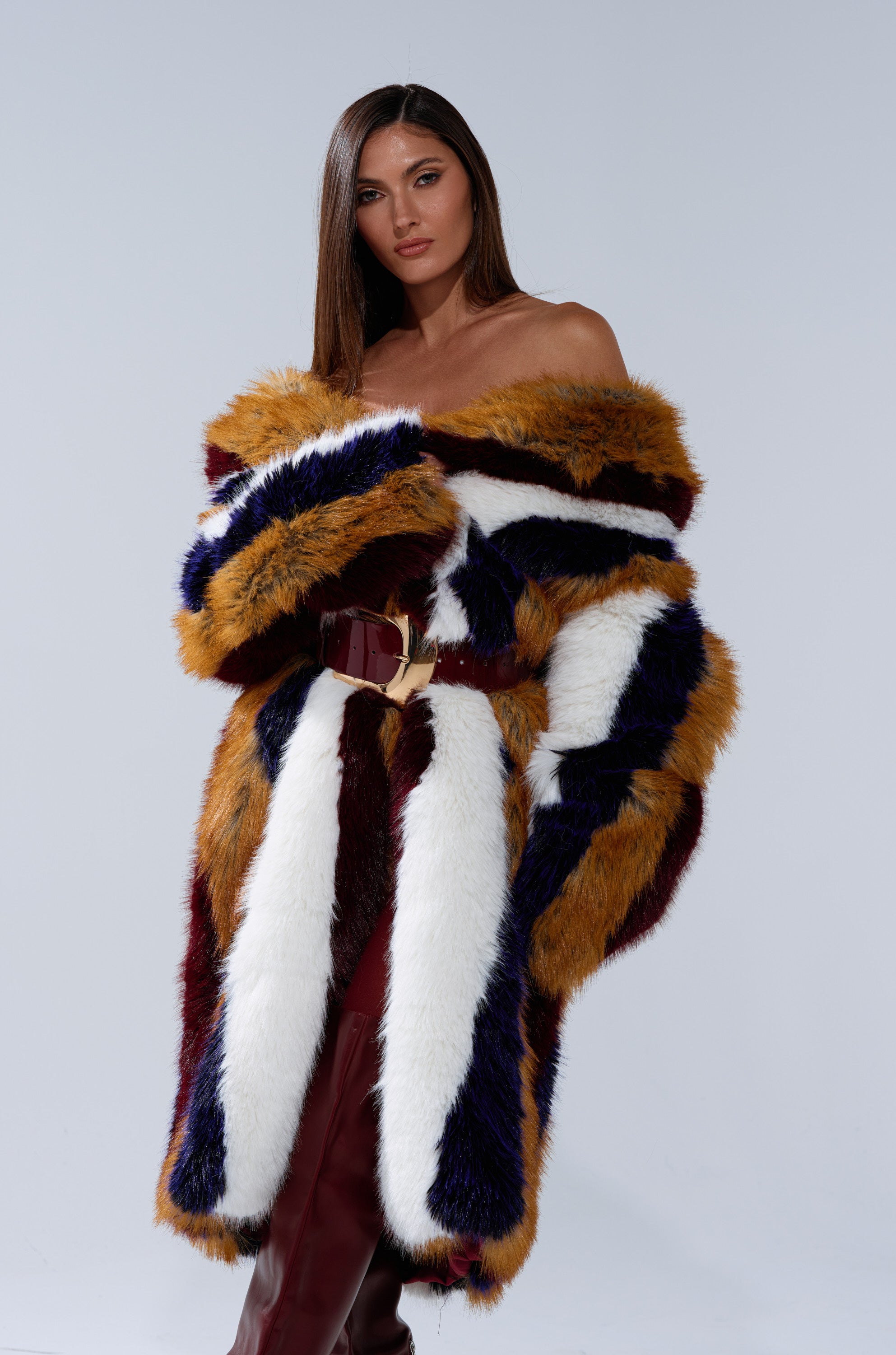 A woman with straight brown hair exudes modern glamor in the ALAIA STRIPED FAUX FUR longline coat, featuring bold white, yellow, navy, and burgundy bands, cinched at the waist against a plain light background.
