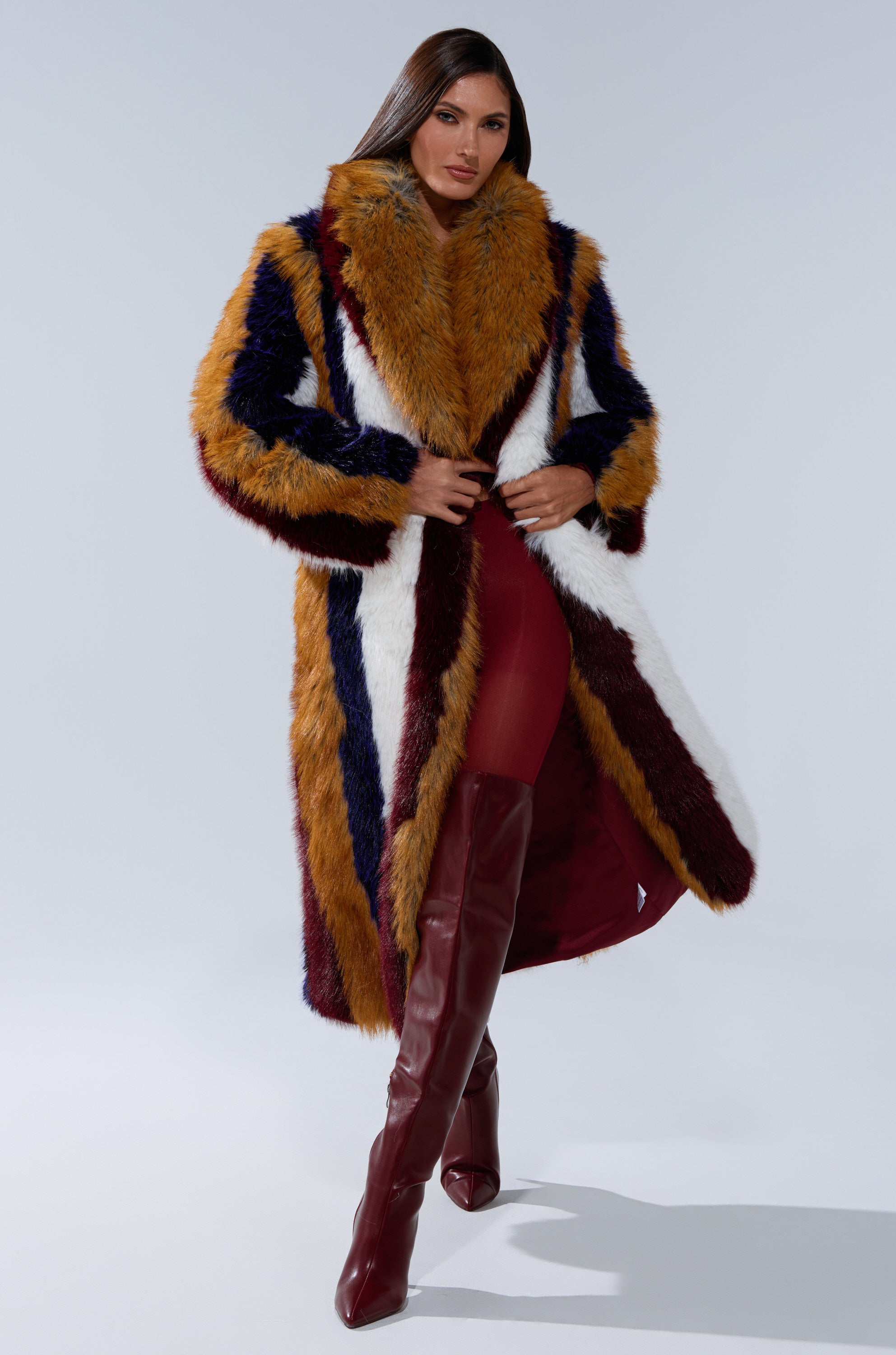 A woman poses confidently in the ALAIA STRIPED FAUX FUR coat, featuring bold gold, burgundy, white, and navy stripes. She pairs it with matching burgundy thigh-high boots and fitted pants against a plain background.