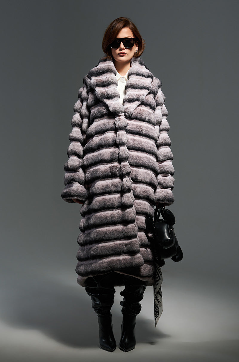 Clad in the LINCOLN PARK FAUX FUR COAT, which boasts a thick, striped design, a person exudes confidence and style against a gray backdrop. They complement the look with dark sunglasses and over-the-knee black boots while holding a black bag elegantly in one hand.