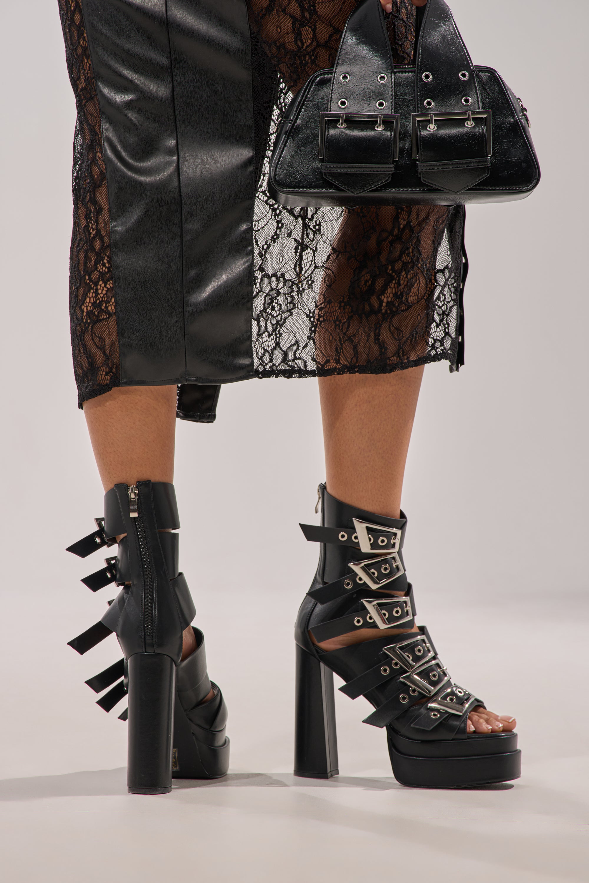 A person wears a black lace and leather skirt, holds a matching black handbag with buckles, and sports AKIRA GOLD ALBERIC BLACK CHUNKY PLATFORM SANDALS. The background is plain and light-colored. 0