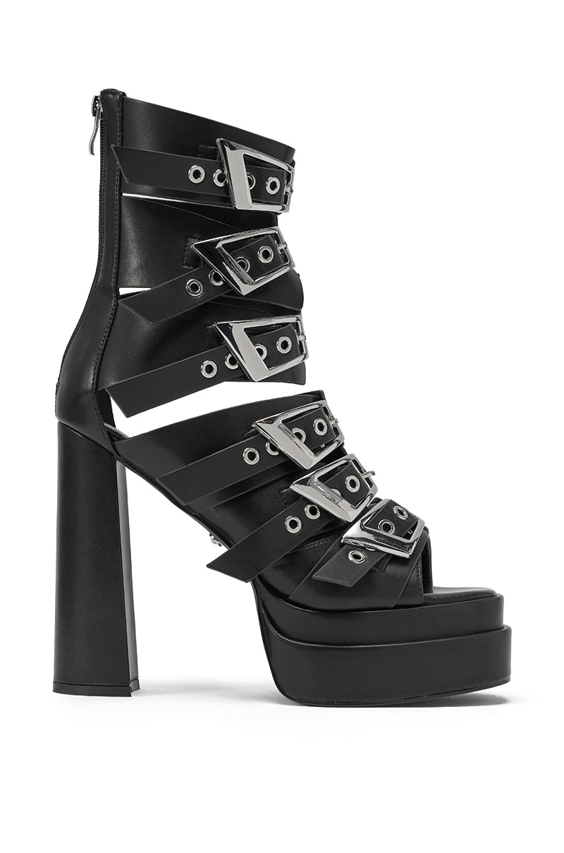 The AKIRA GOLD ALBERIC BLACK CHUNKY PLATFORM SANDAL is a high-heeled black faux leather sandal with wide straps, silver grommets, bold buckles, a chunky block heel, and a back zip closure. 2