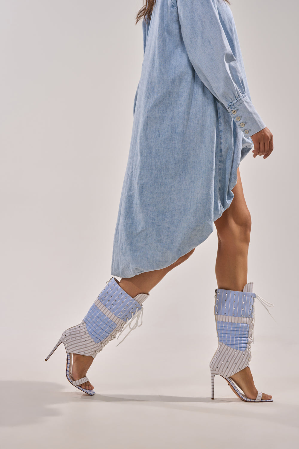 A woman in a light blue oversized shirt dress and AKIRA Gold Alberto Blue Stripe Pattern Gladiator Stiletto heels with knee-high lace-up details walks across a plain background. 1