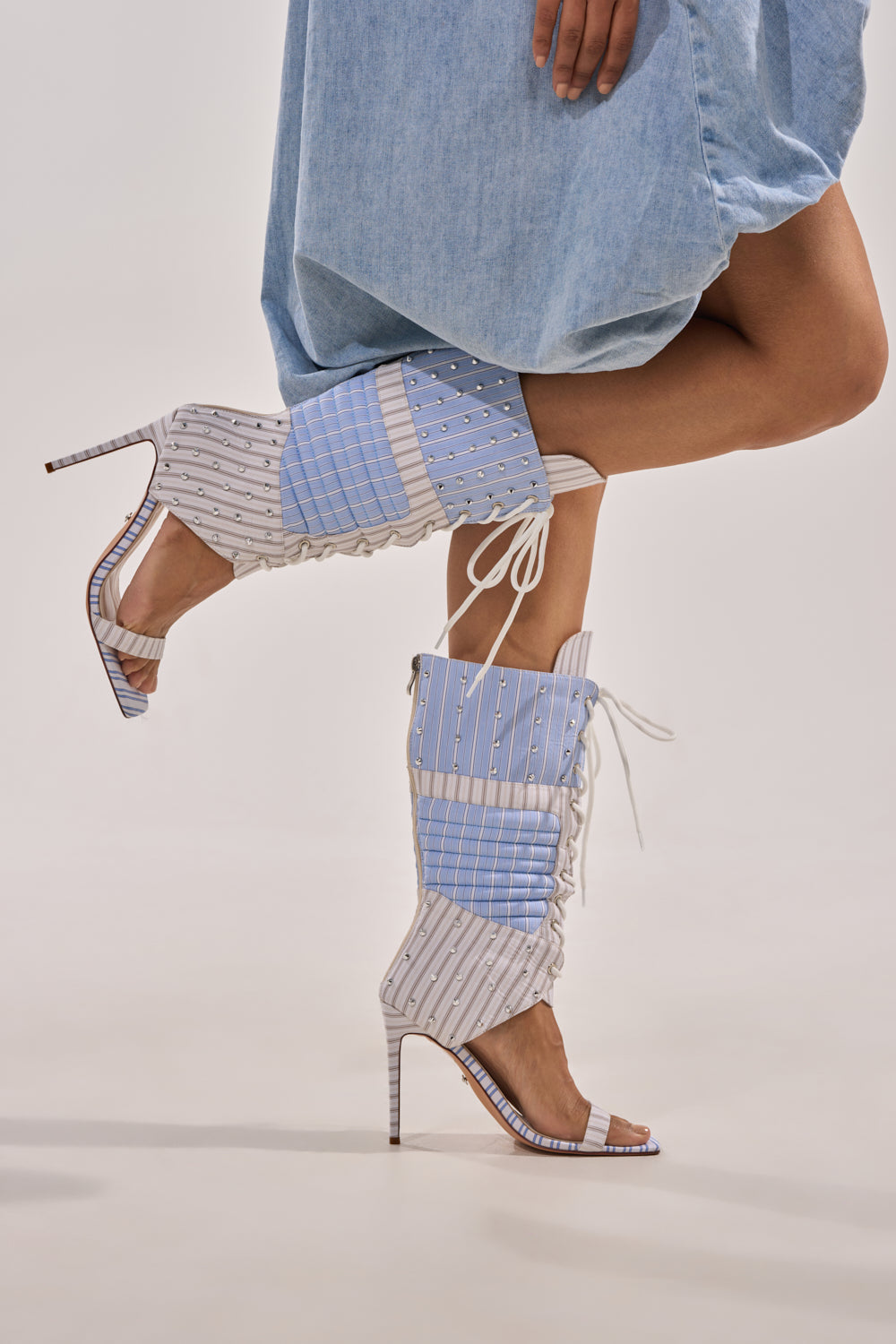 A person poses fashionably, wearing a light blue, knee-length skirt paired with the AKIRA GOLD ALBERTO BLUE STRIPE PATTERN GLADIATOR STILETTO—mid-calf boots featuring blue and white stripes, lace-up details, and thin high heels. 7