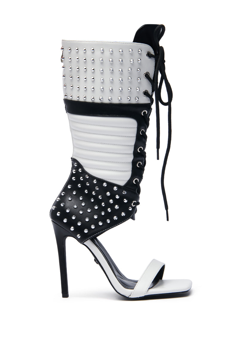 The AZALEA WANG ALBERTO BLACK WHITE STUDDED OPEN TOE BOOT features a high stiletto heel, open-toe design, and faux leather construction. Adorned with silver studs, lace-up sides, and a quilted middle section, it seamlessly merges sandal and boot elements.